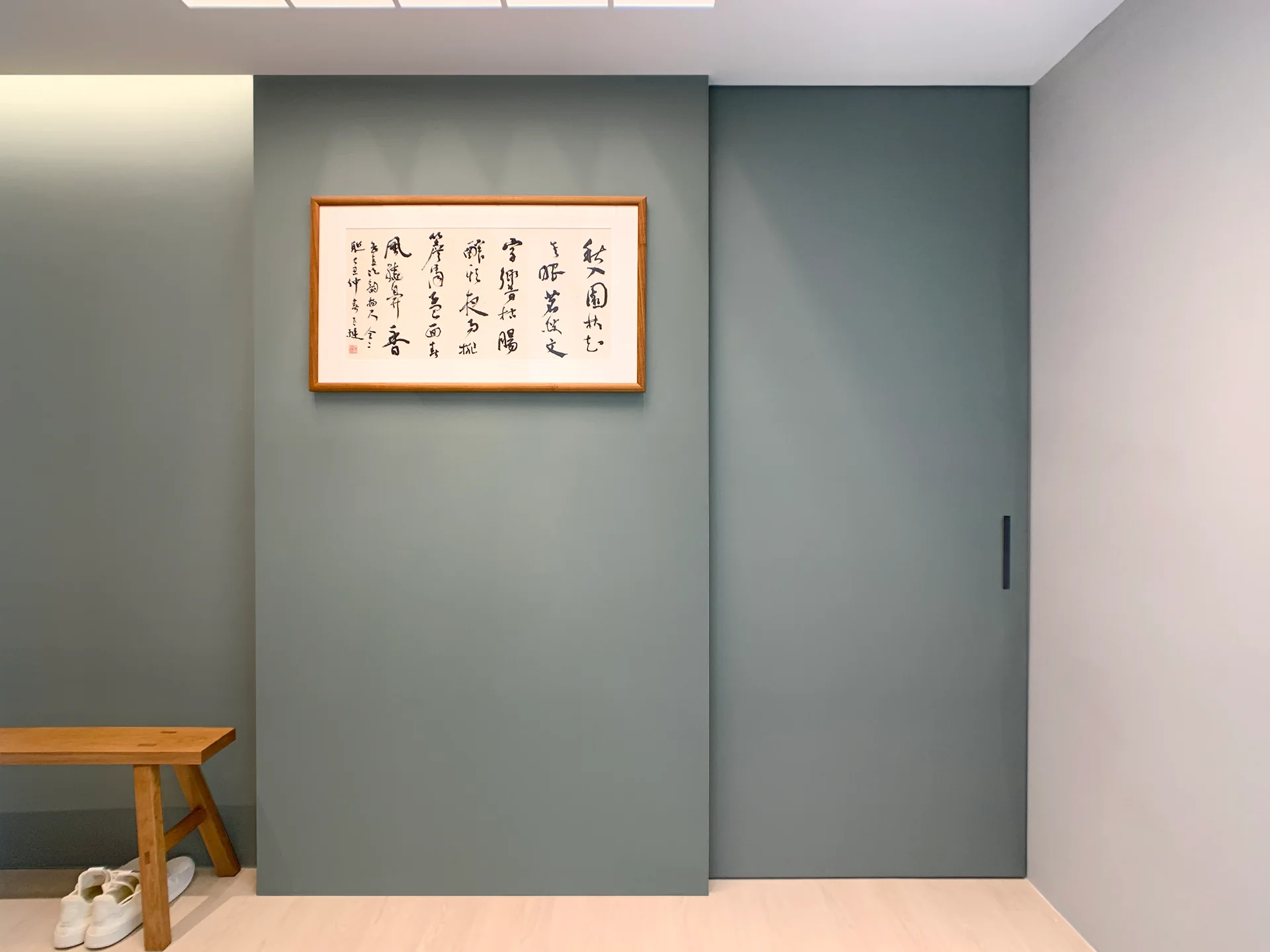 A minimalist entryway features a blue-grey accent wall with a framed calligraphy art piece, a matching sliding door, and a wooden bench.