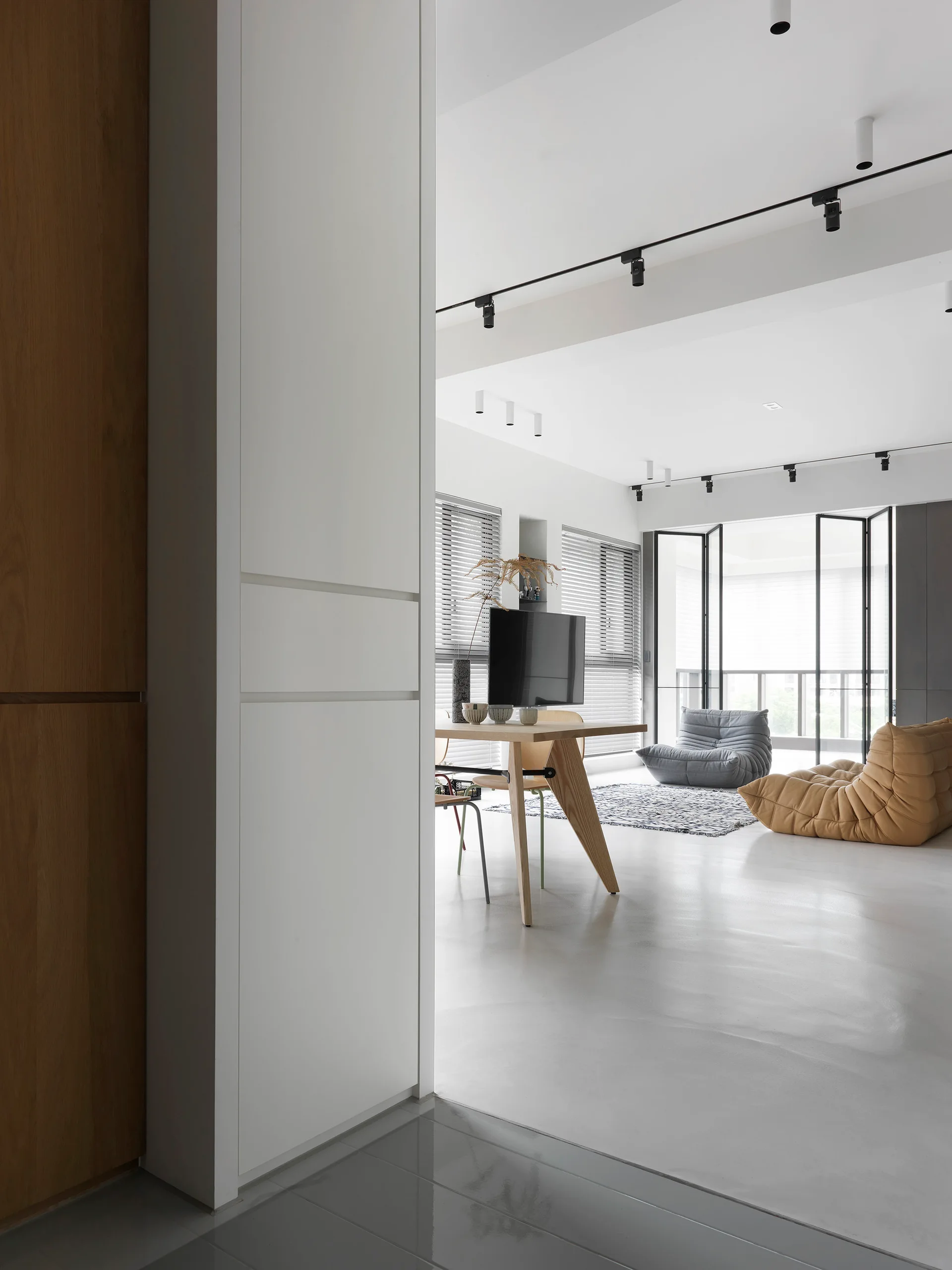 A bright minimalist living room features a wooden dining table, black ceiling track lights, and two slouchy low-profile lounge chairs on a polished floor.