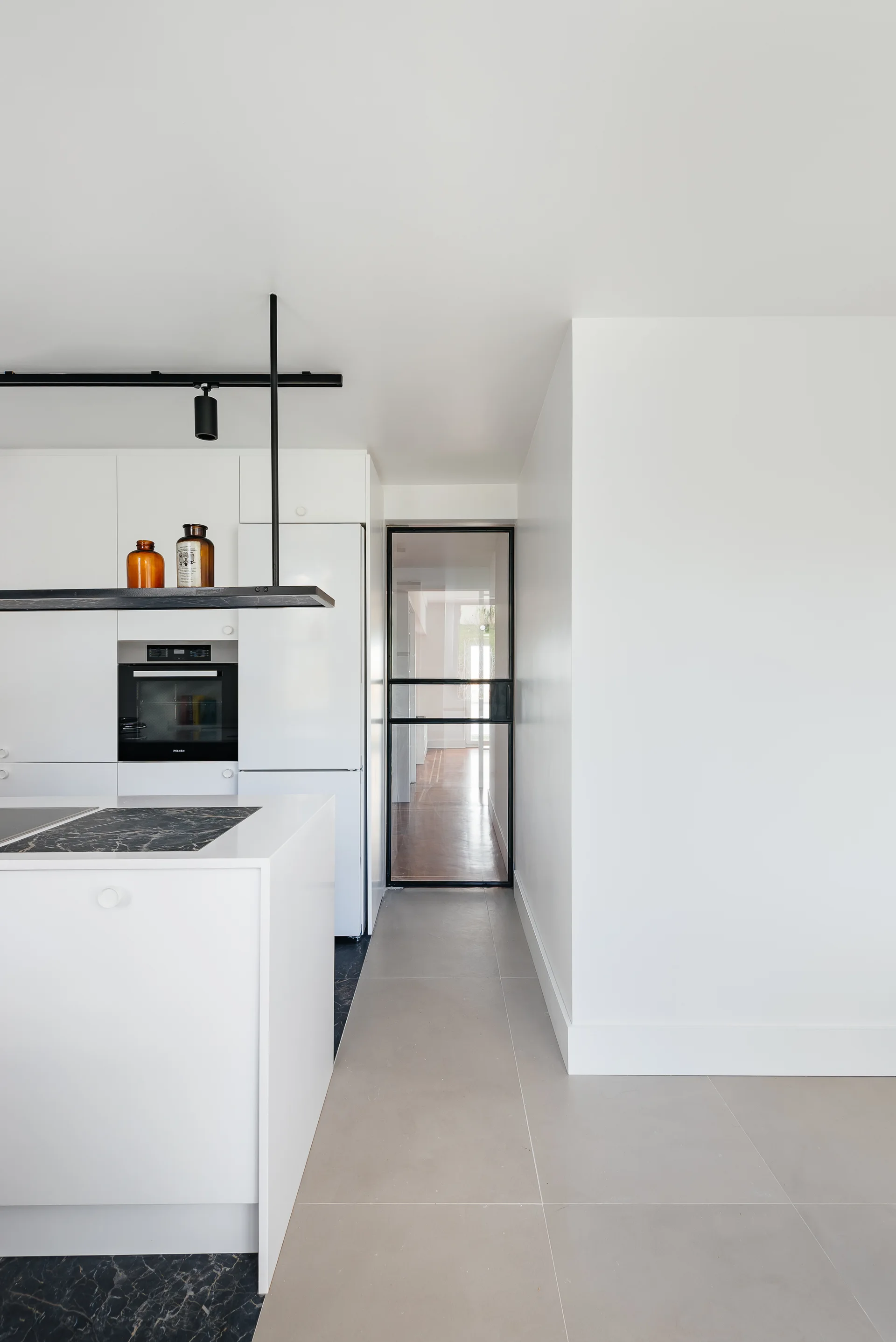 A minimalist white kitchen features flat-panel cabinets, a black stone island countertop, and a black-framed glass door leading to a hallway.