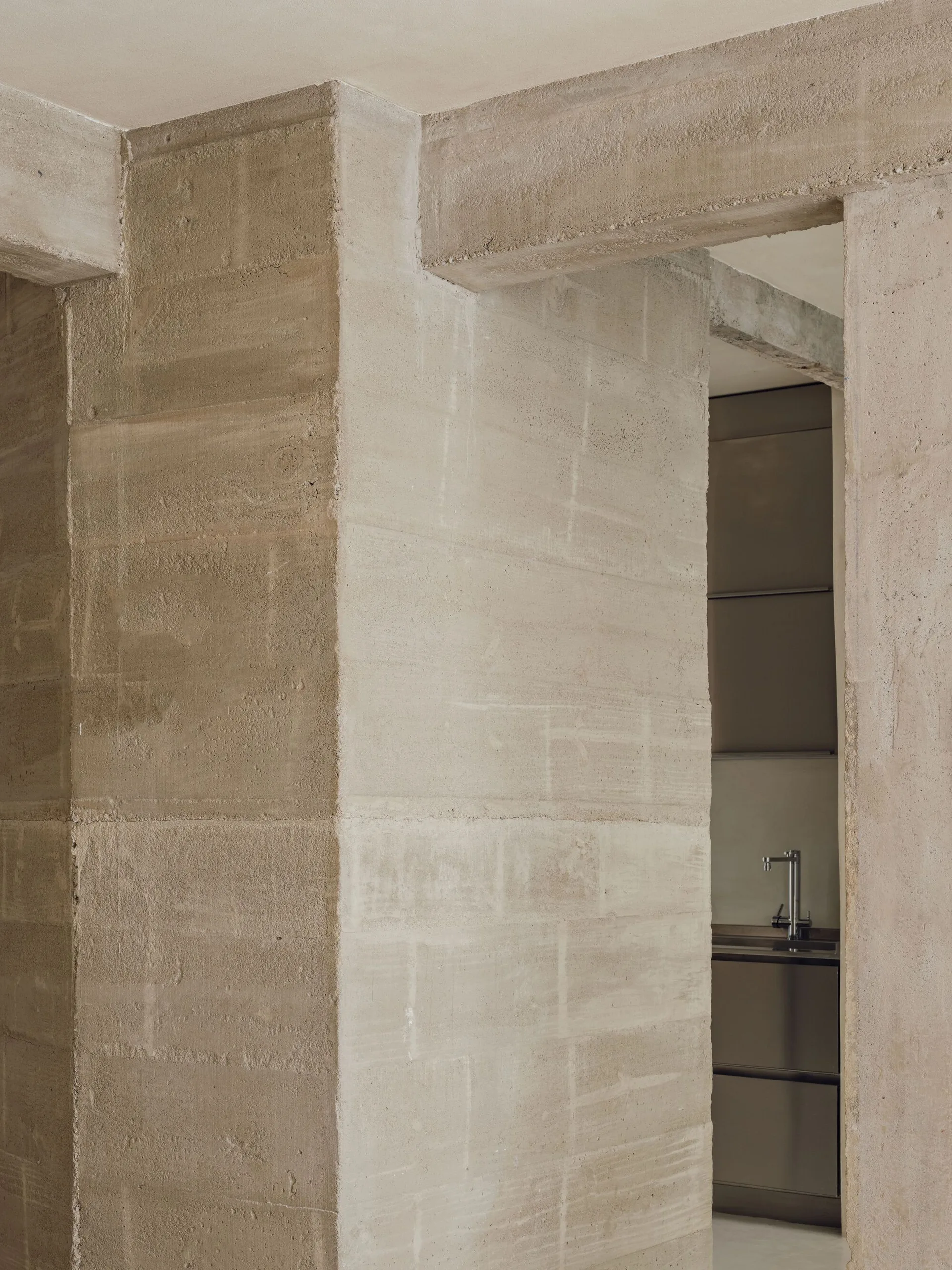 A view through a rough, textured concrete archway reveals a minimalist modern kitchen with sleek grey cabinets and a silver faucet.
