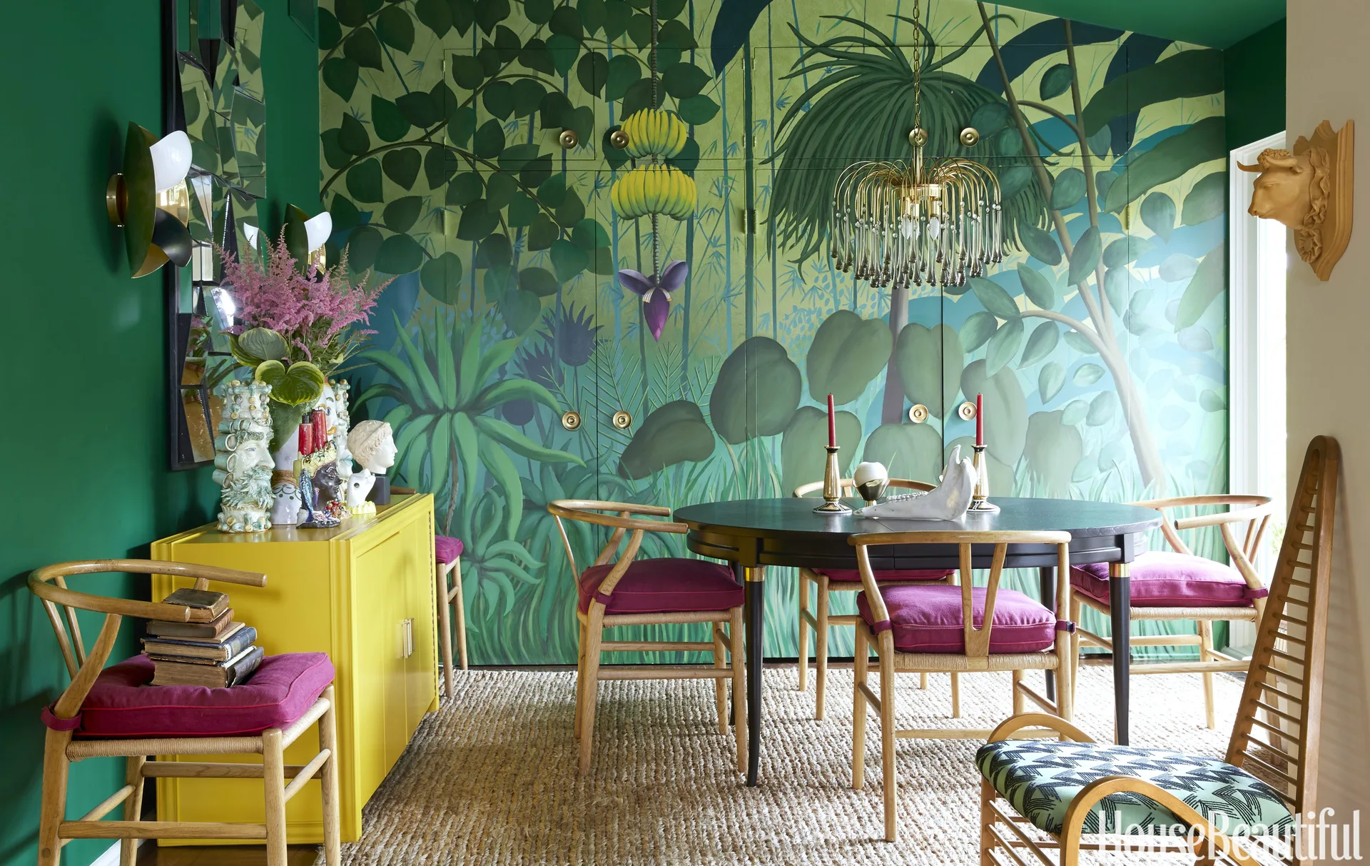 A colorful green dining room features a large jungle mural, a black circular table with magenta-cushioned chairs, and a bright yellow sideboard.