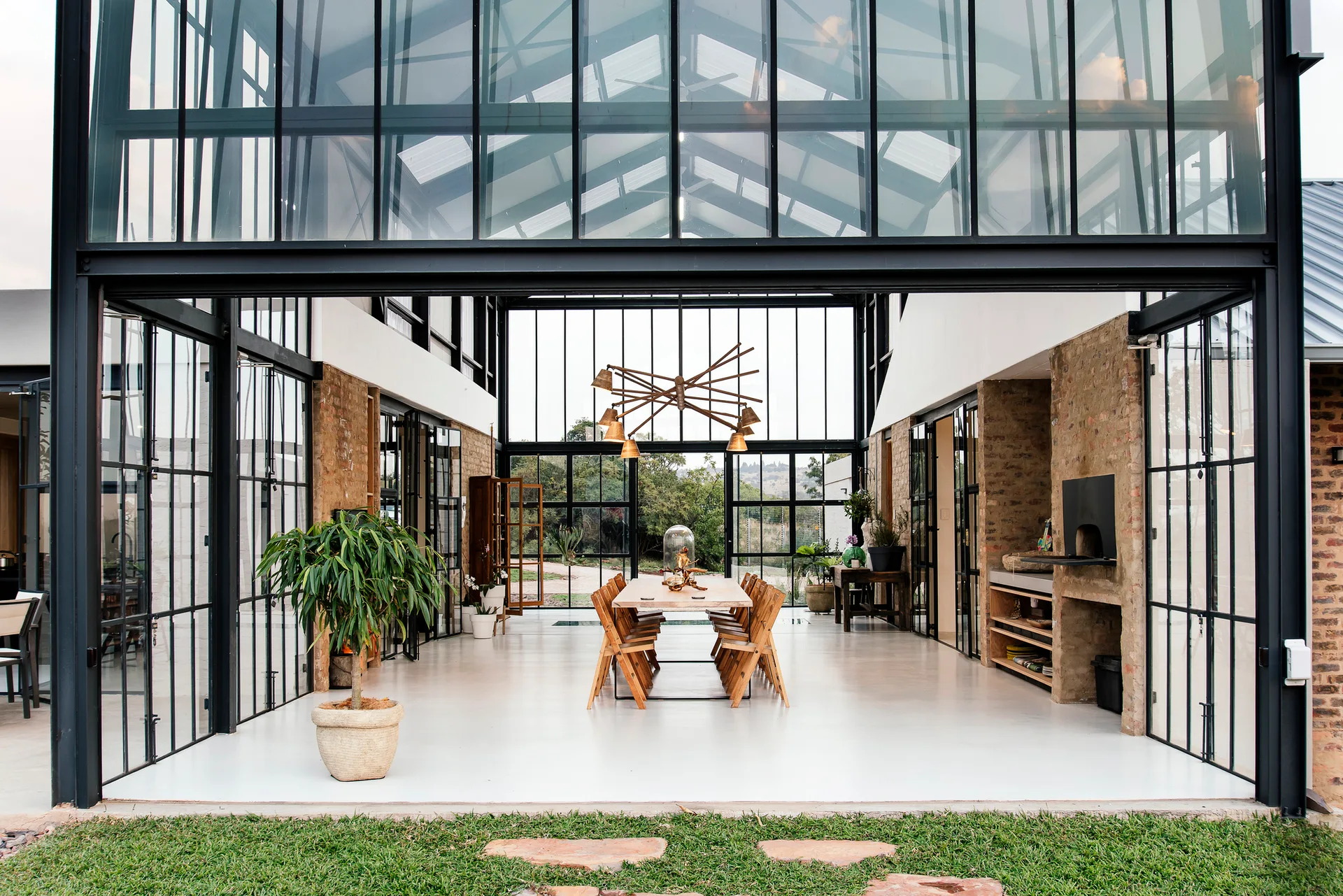 An open dining pavilion with a glass roof, a long wooden table, black metal window frames, exposed brick pillars, and white floors.