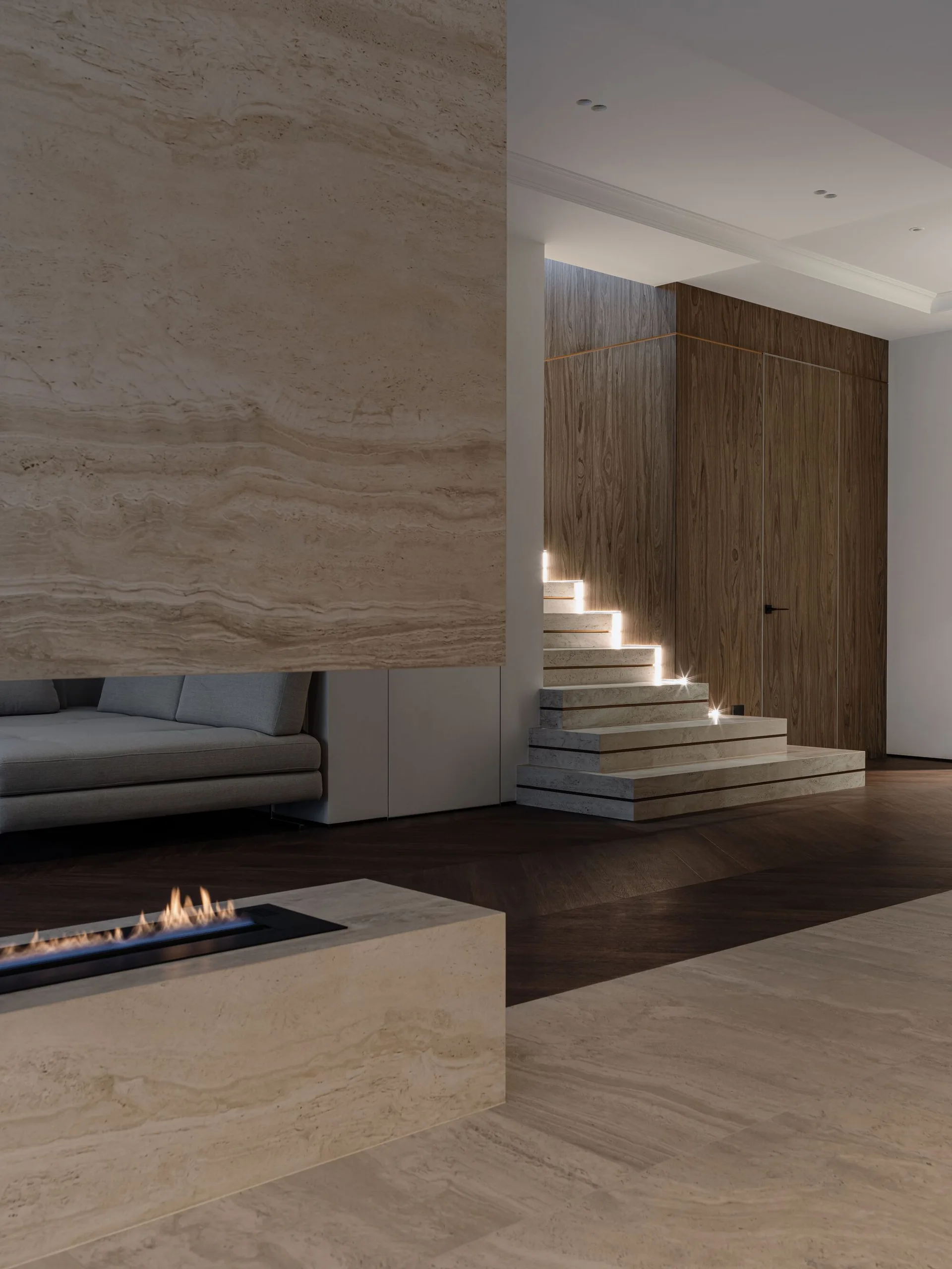 A modern interior featuring a beige travertine fireplace, dark wood herringbone flooring, and a stone staircase with integrated LED step lighting.