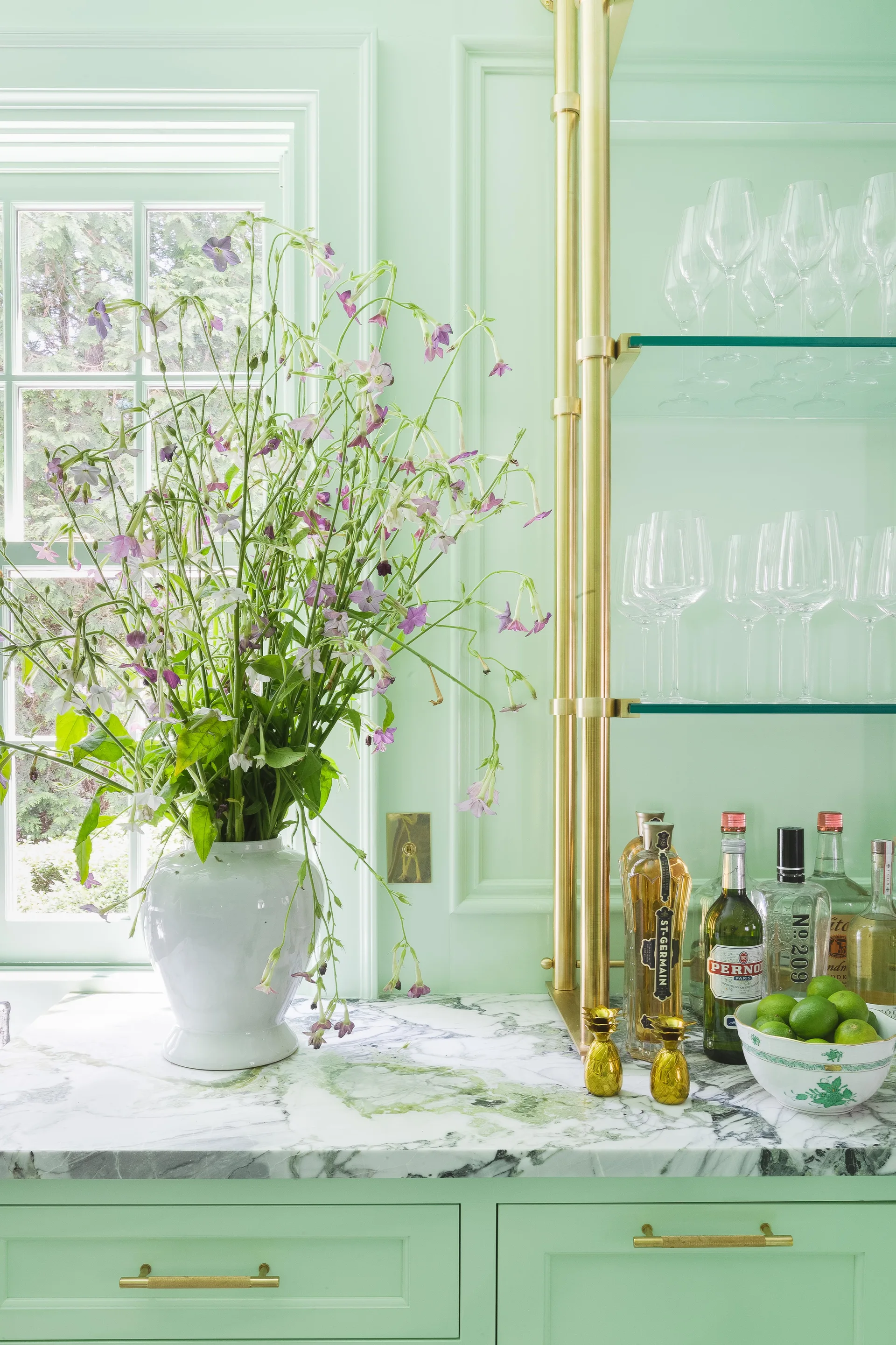 A mint green home bar features a green-veined marble countertop, brass shelving with glassware, and a white vase with purple flowers.