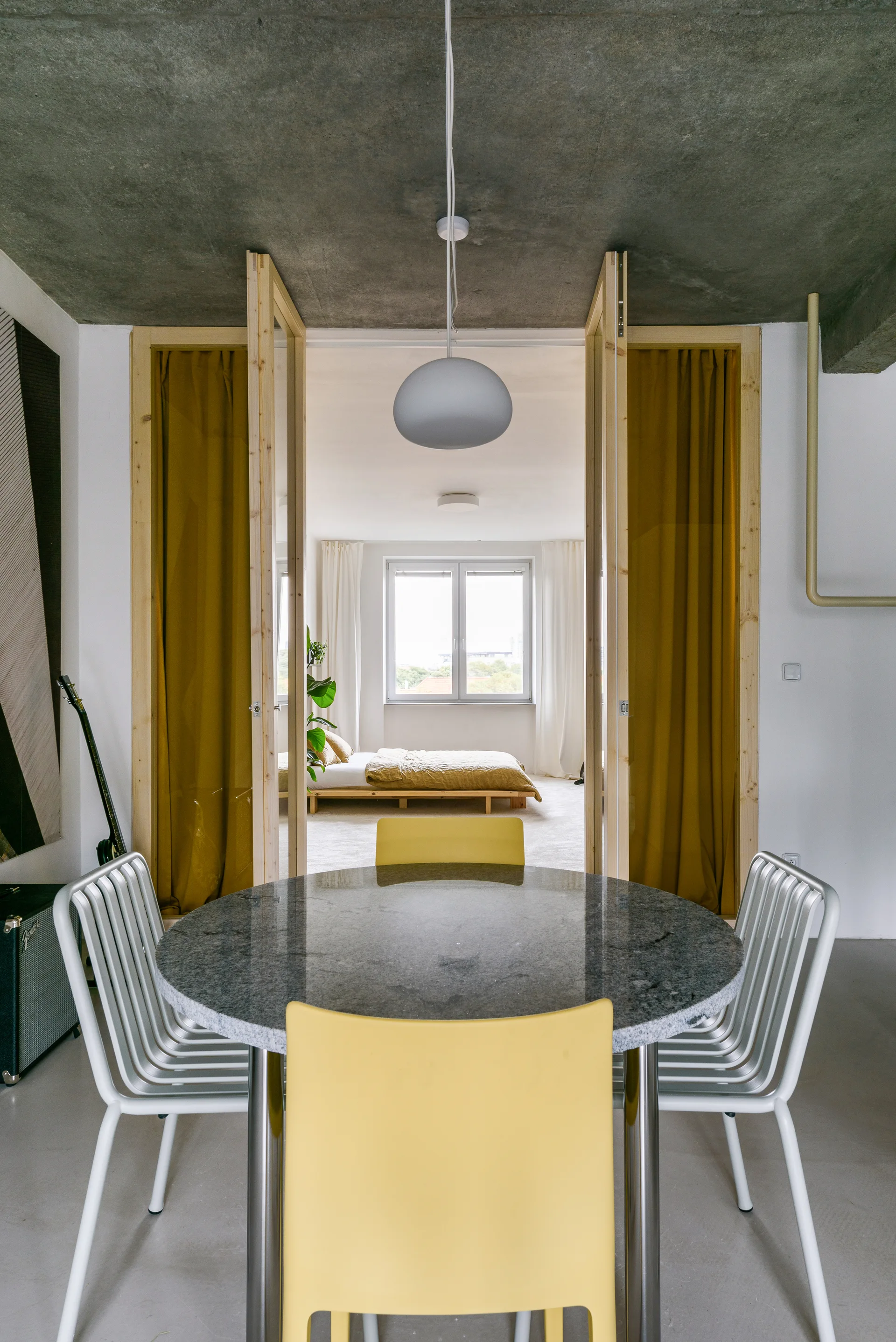 A round grey stone dining table surrounded by yellow and white metal chairs sits under a concrete ceiling, looking into a bedroom with mustard yellow curtains.