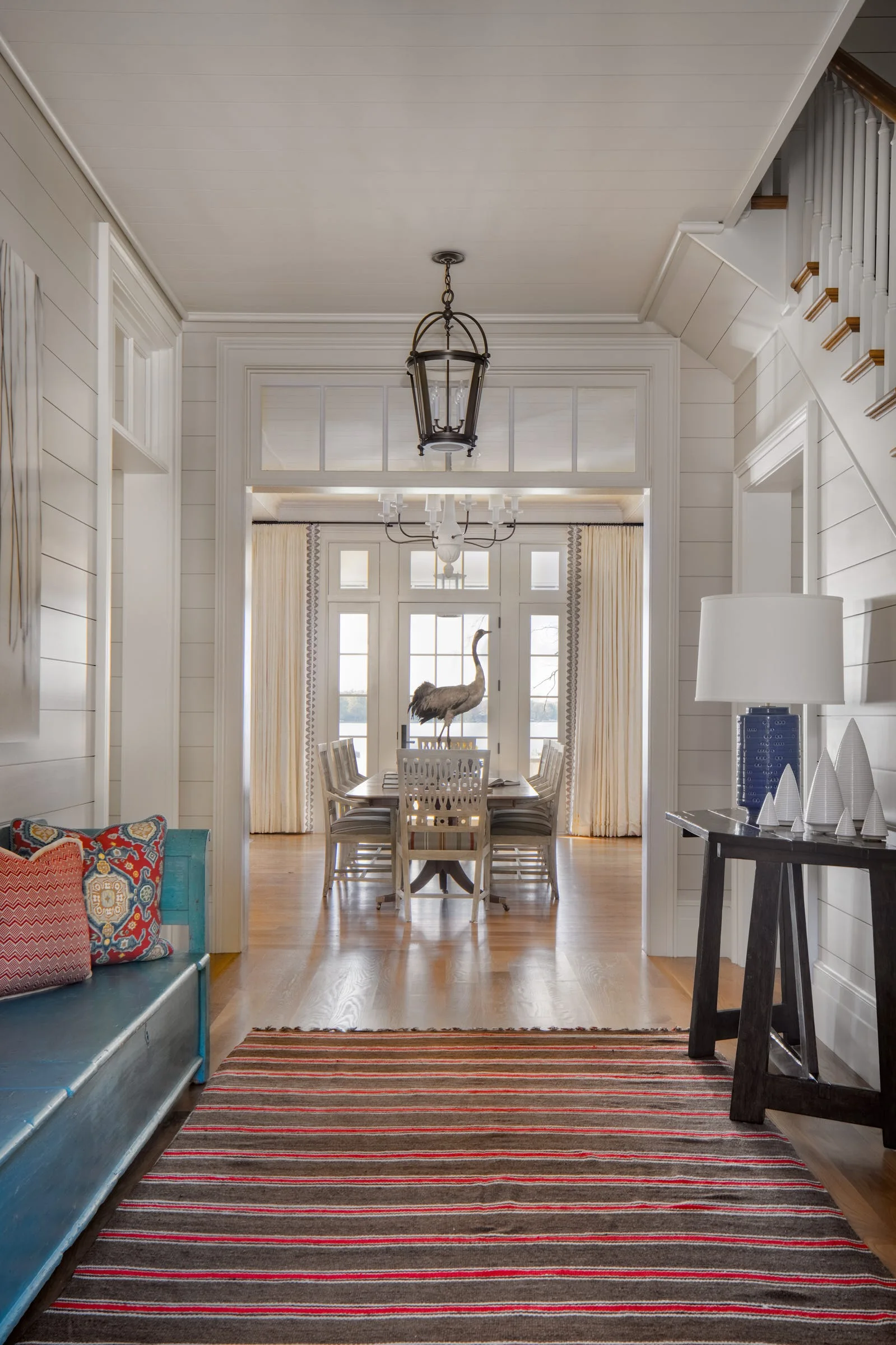 An entryway with white shiplap walls, a blue bench, and a striped rug leads to a dining room seen through an open doorway.