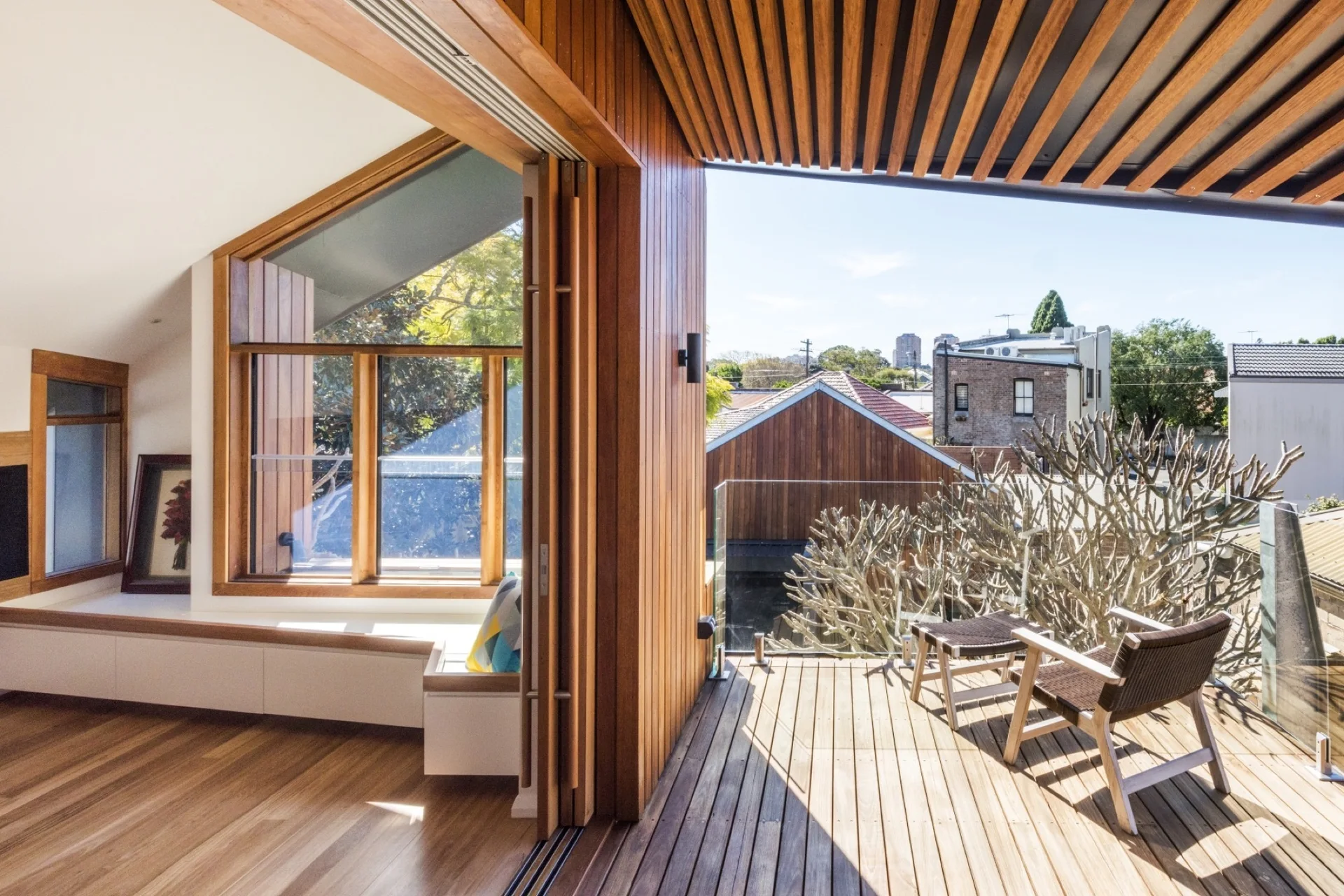 A contemporary wooden balcony with two chairs and a glass railing connects to an indoor living area with a built-in window seat.