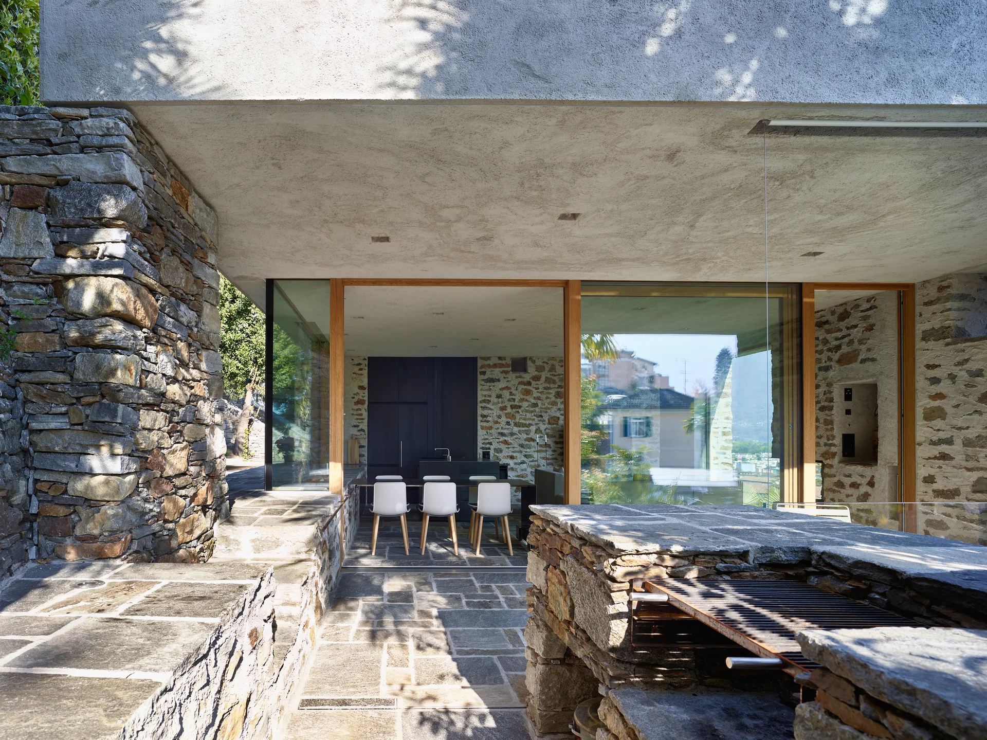 A rustic stone patio features a built-in stone grill and large sliding glass doors opening to an indoor dining area with white chairs.