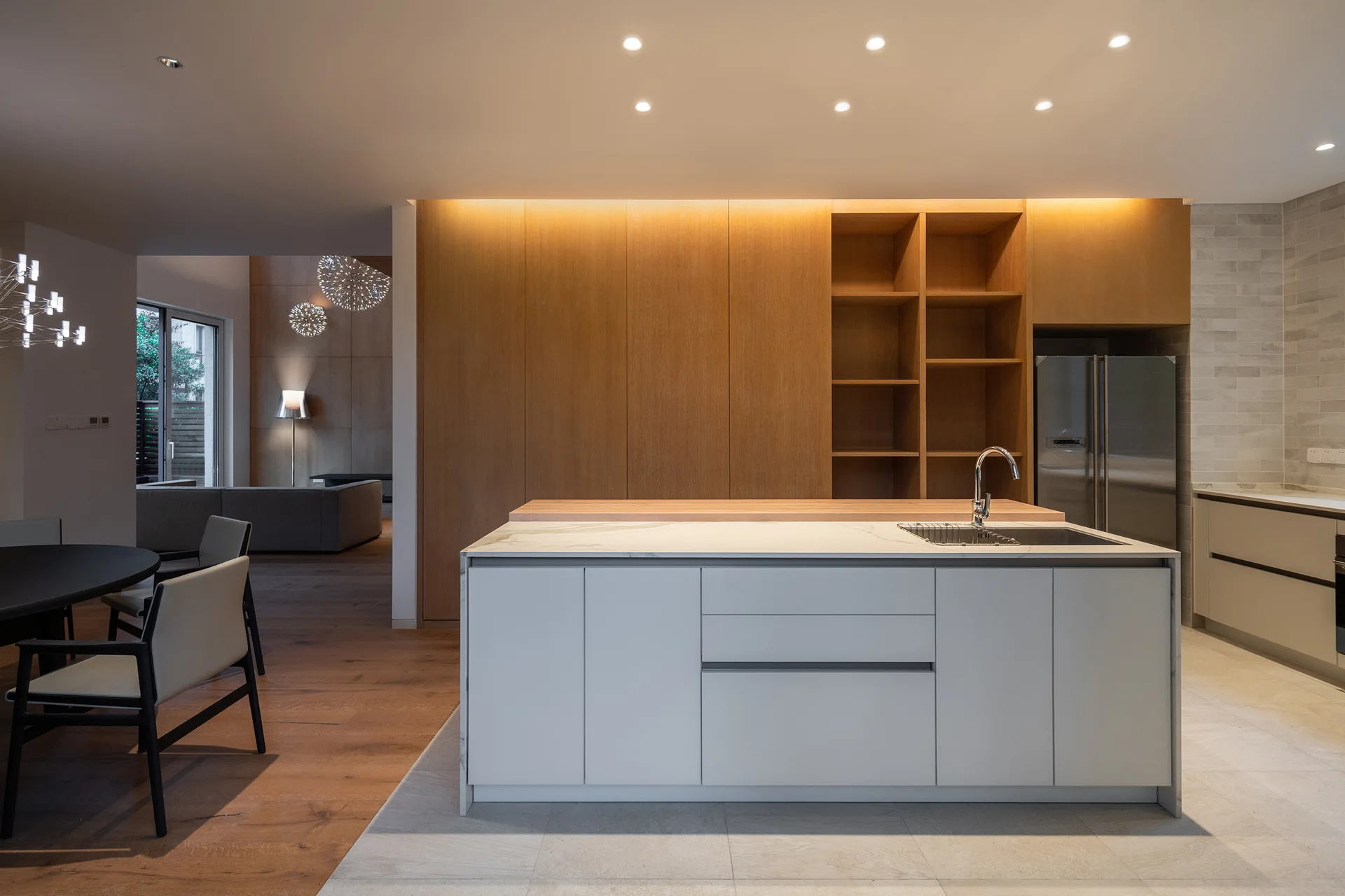 A modern open kitchen features a white stone island with a sink, backed by a wall of floor-to-ceiling wood cabinets under soft cove lighting.