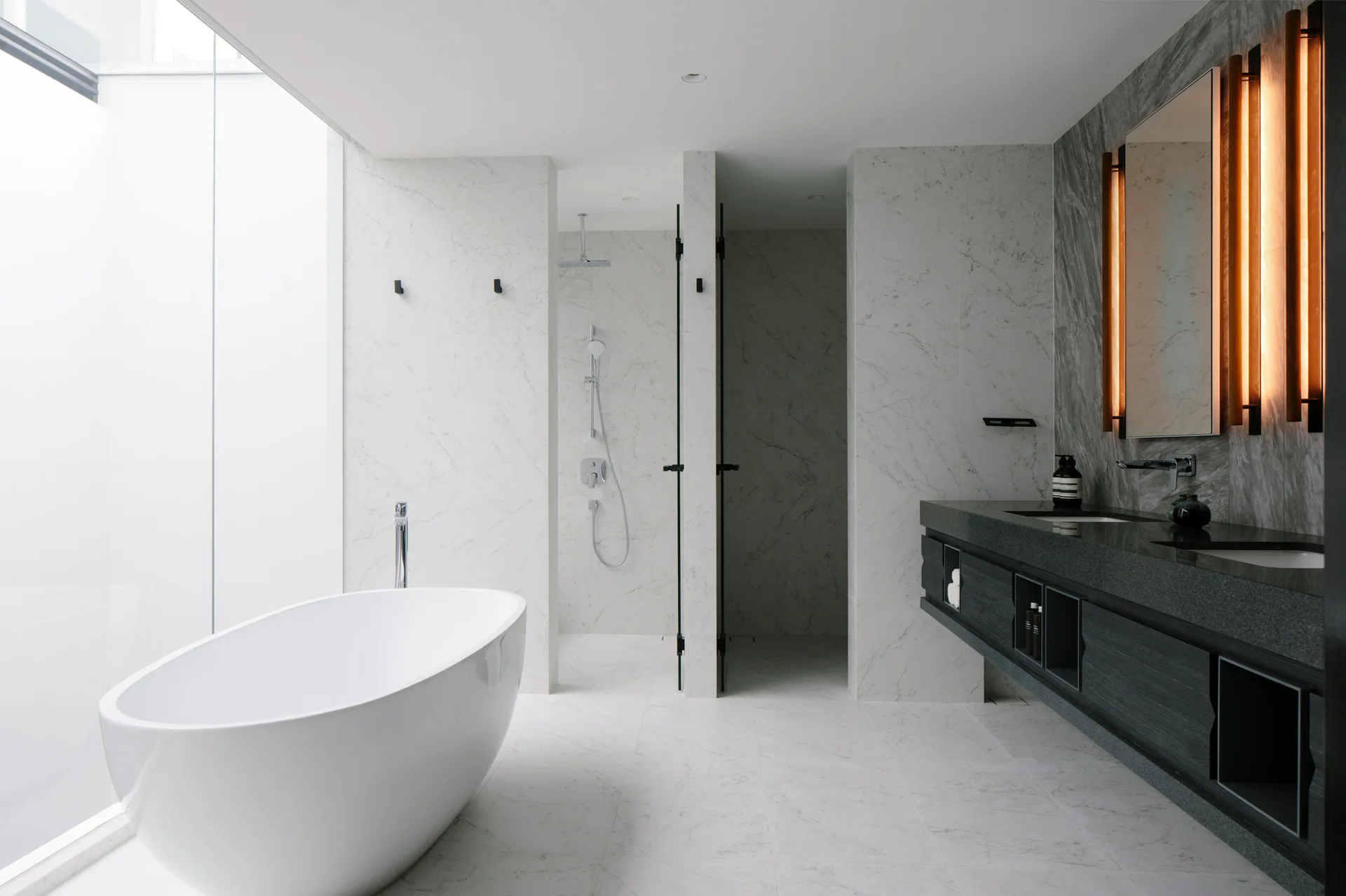 A modern white marble bathroom features a freestanding tub, a walk-in shower, and a dark floating vanity with glowing vertical mirror lights.