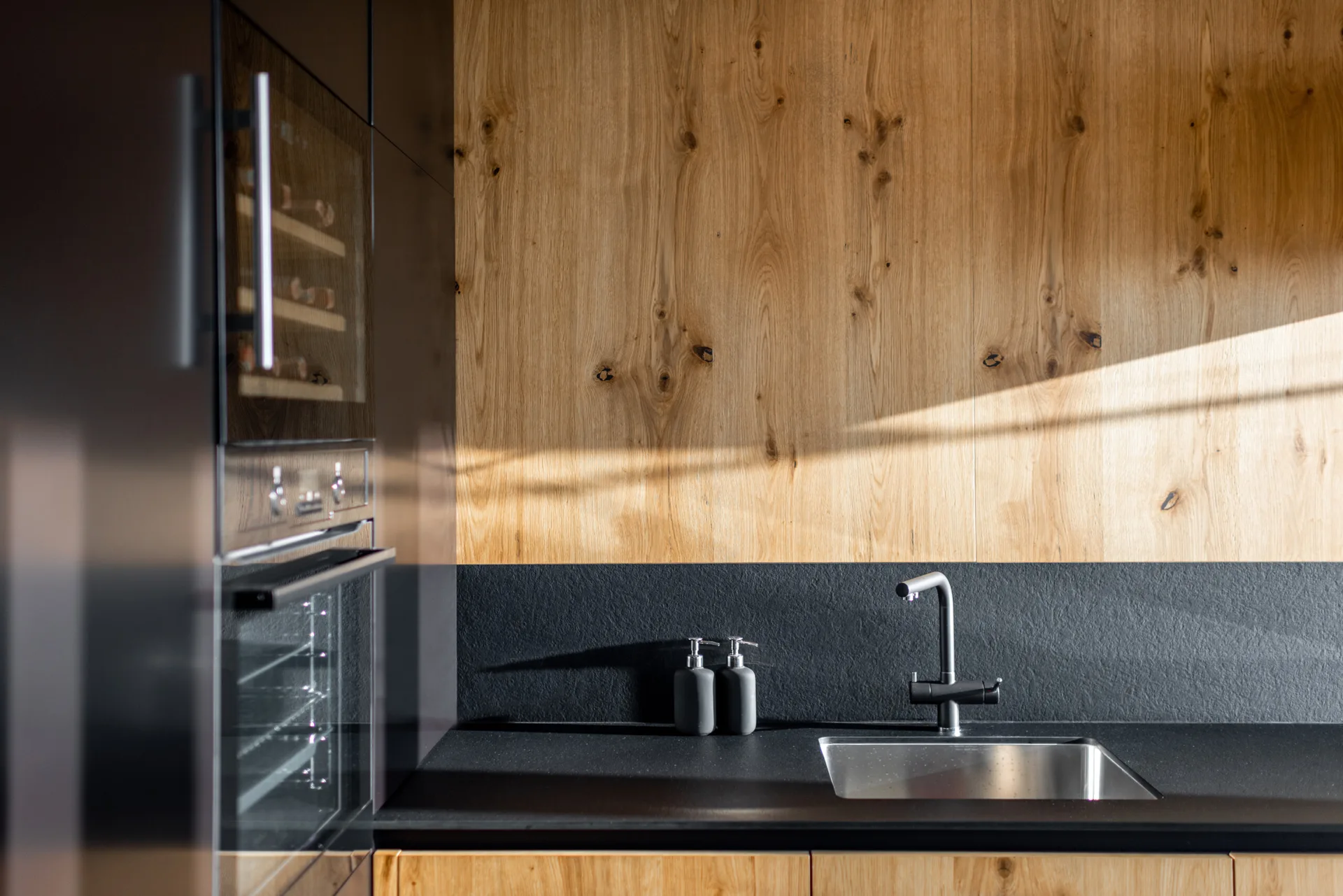 A modern kitchen sink area features a stainless steel sink, matte black countertops, a dark backsplash, and a warm wood paneled wall.
