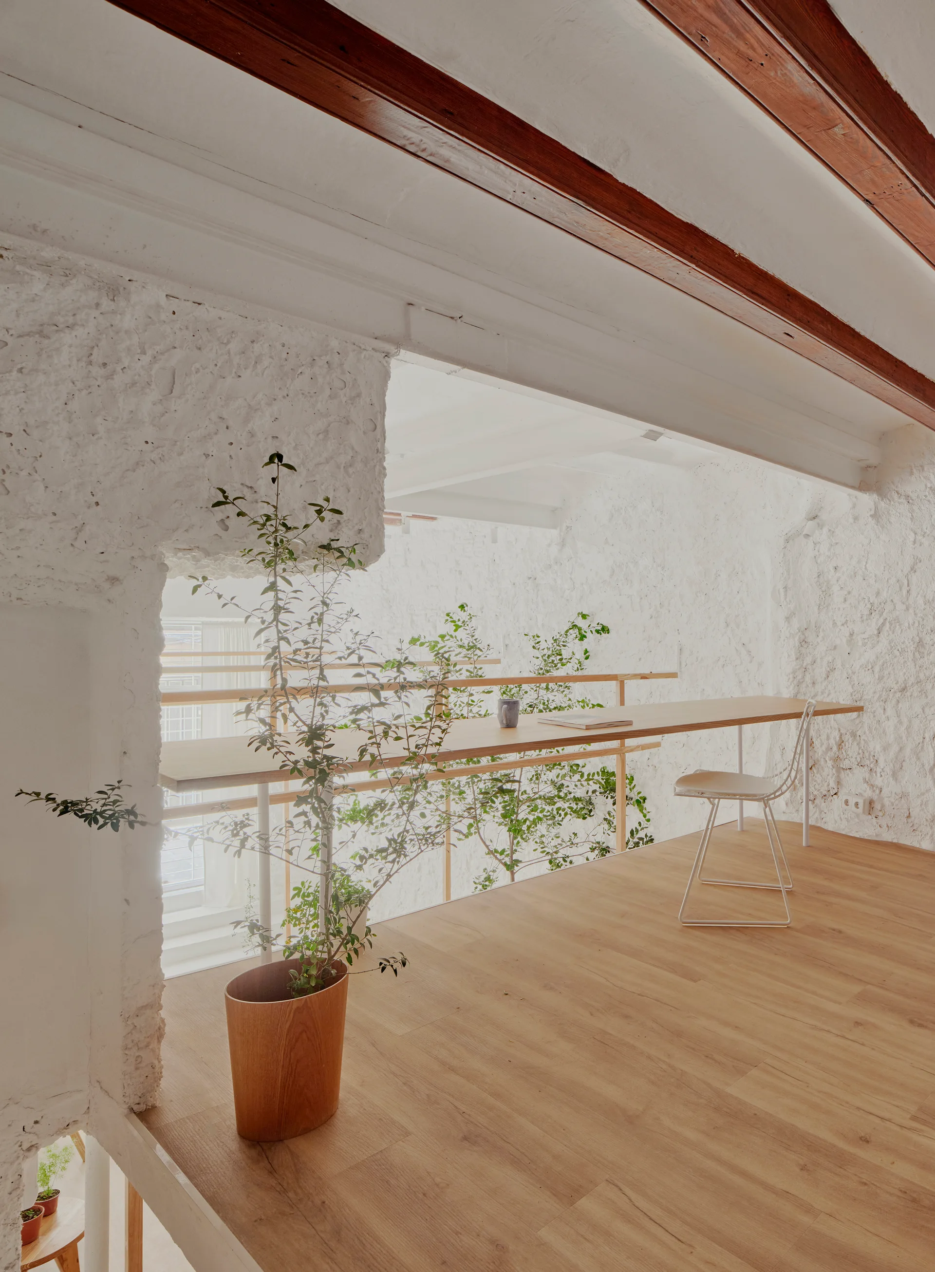 A loft workspace features a long wood desk, a white wire chair, and a potted plant against rough white plaster walls under exposed ceiling beams.