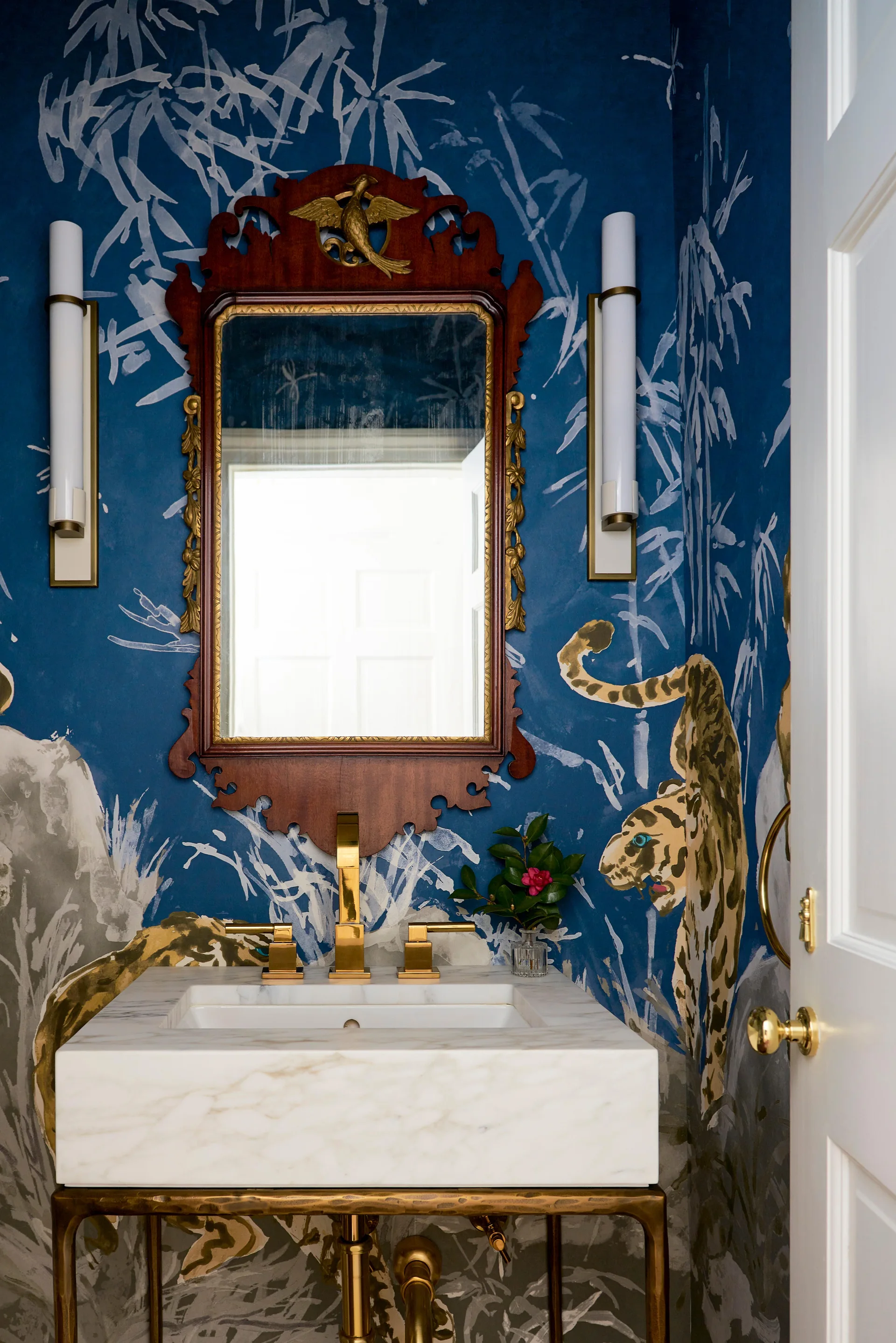 A powder room vanity features a white marble countertop, brass faucet, and an ornate wood mirror set against deep blue tiger and bamboo mural wallpaper.