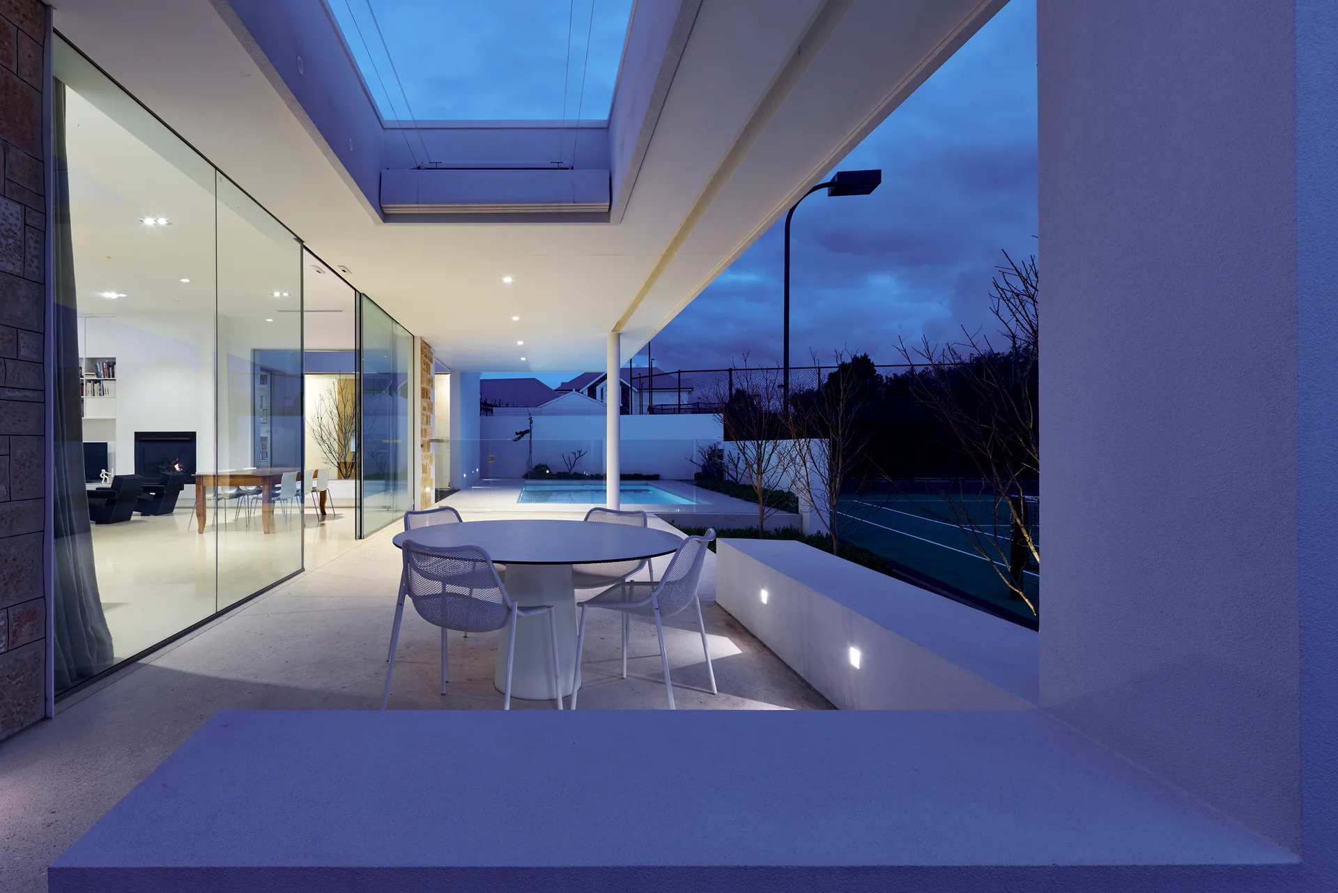 A modern covered patio features a circular white dining set next to a low concrete wall with integrated lights under a twilight sky.