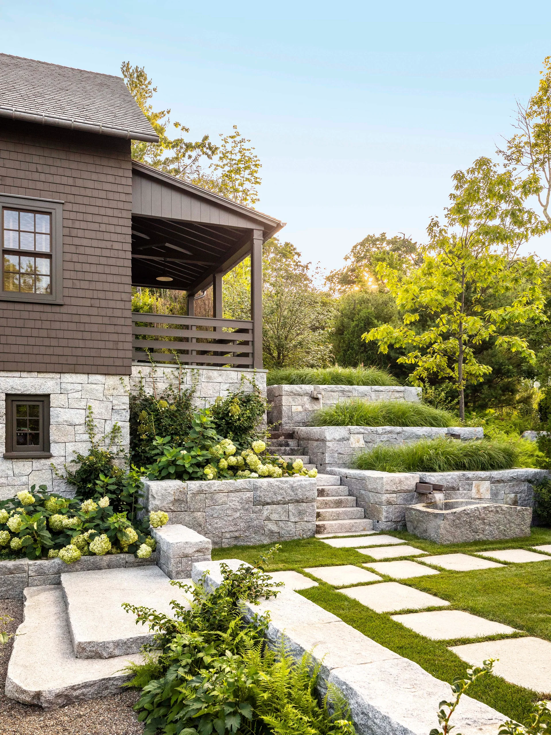 A rustic house with dark brown shingle siding features a tiered stone landscape with stairs, ornamental grasses, and rectangular stone pavers.