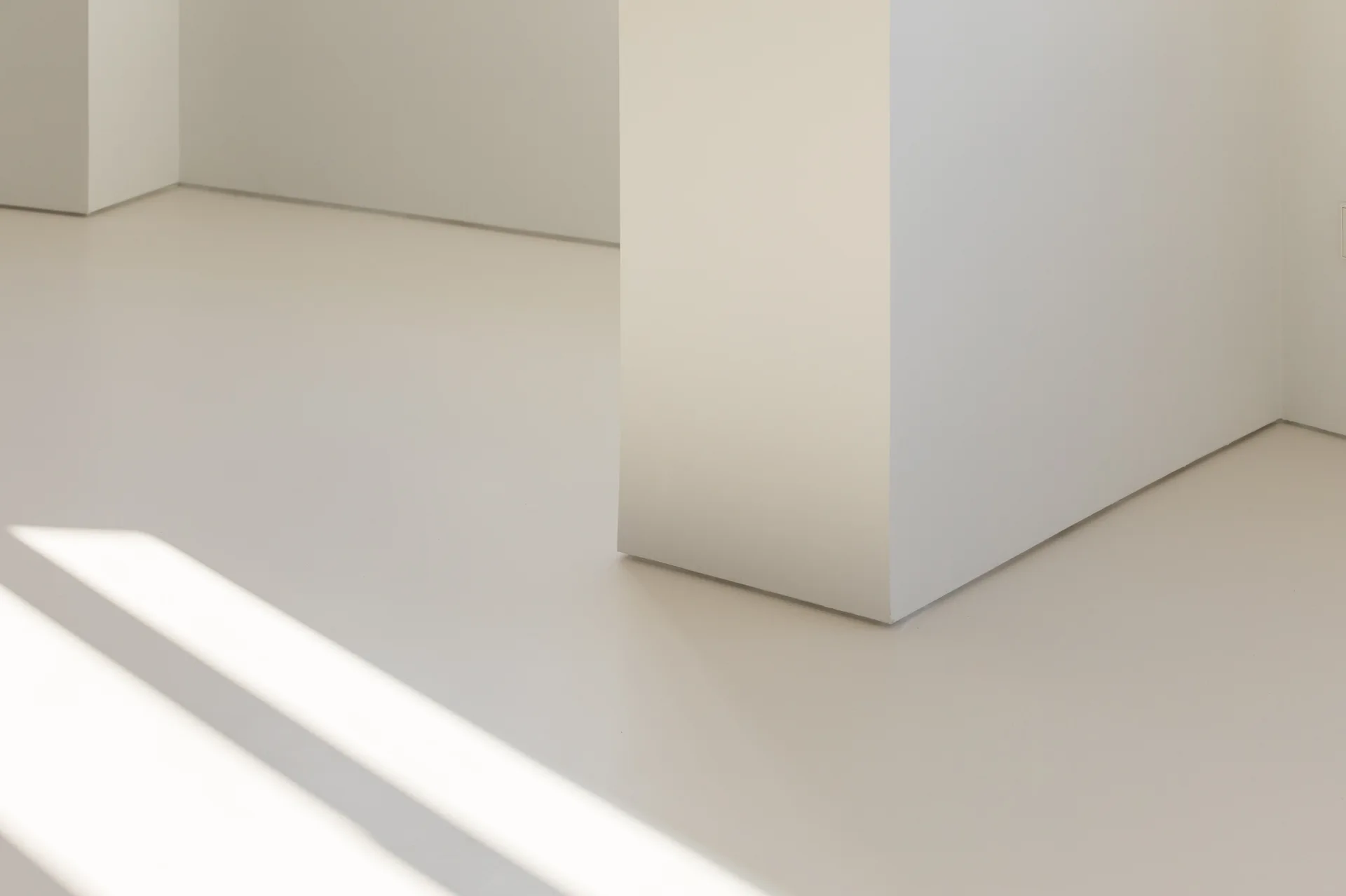 A close-up of a minimalist interior showing the seamless, trim-free intersection of a white matte plaster column and a smooth off-white floor.