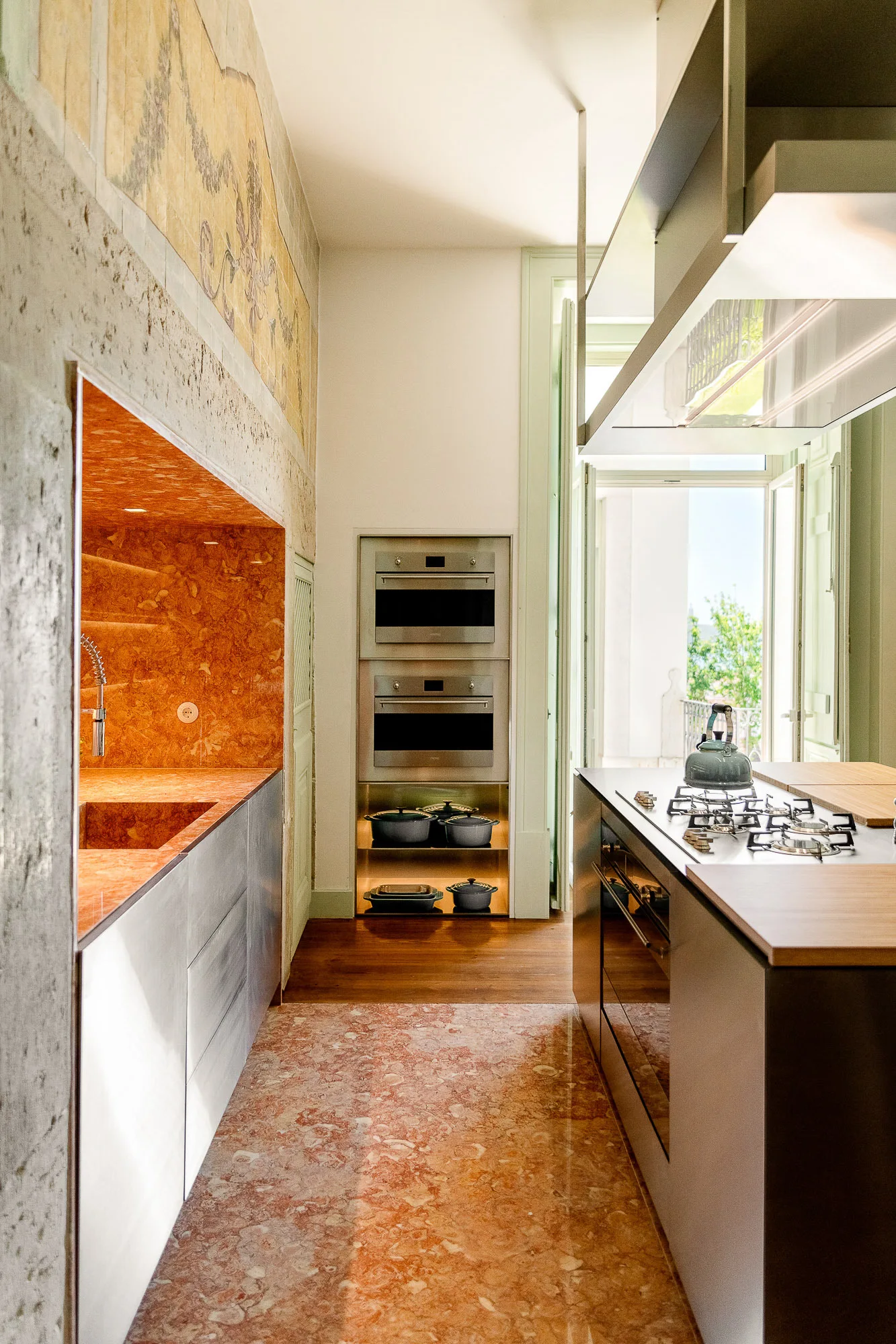 A modern kitchen with a recessed orange marble sink alcove, stainless steel cabinetry, a matching marble floor, and a wall mural.