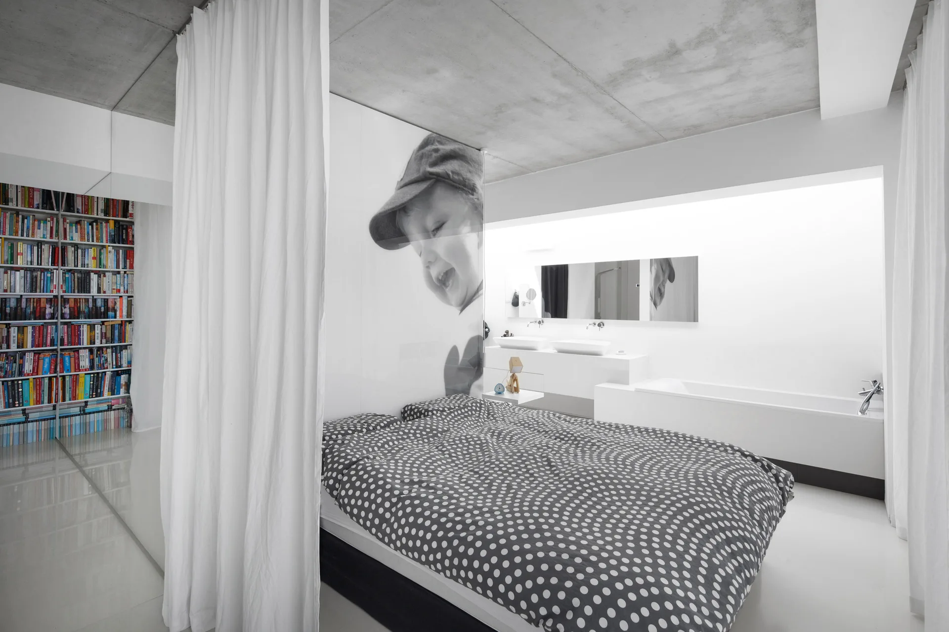 An open-concept bedroom features a bed with polka-dot bedding, an exposed concrete ceiling, and an integrated bathroom with a bathtub.