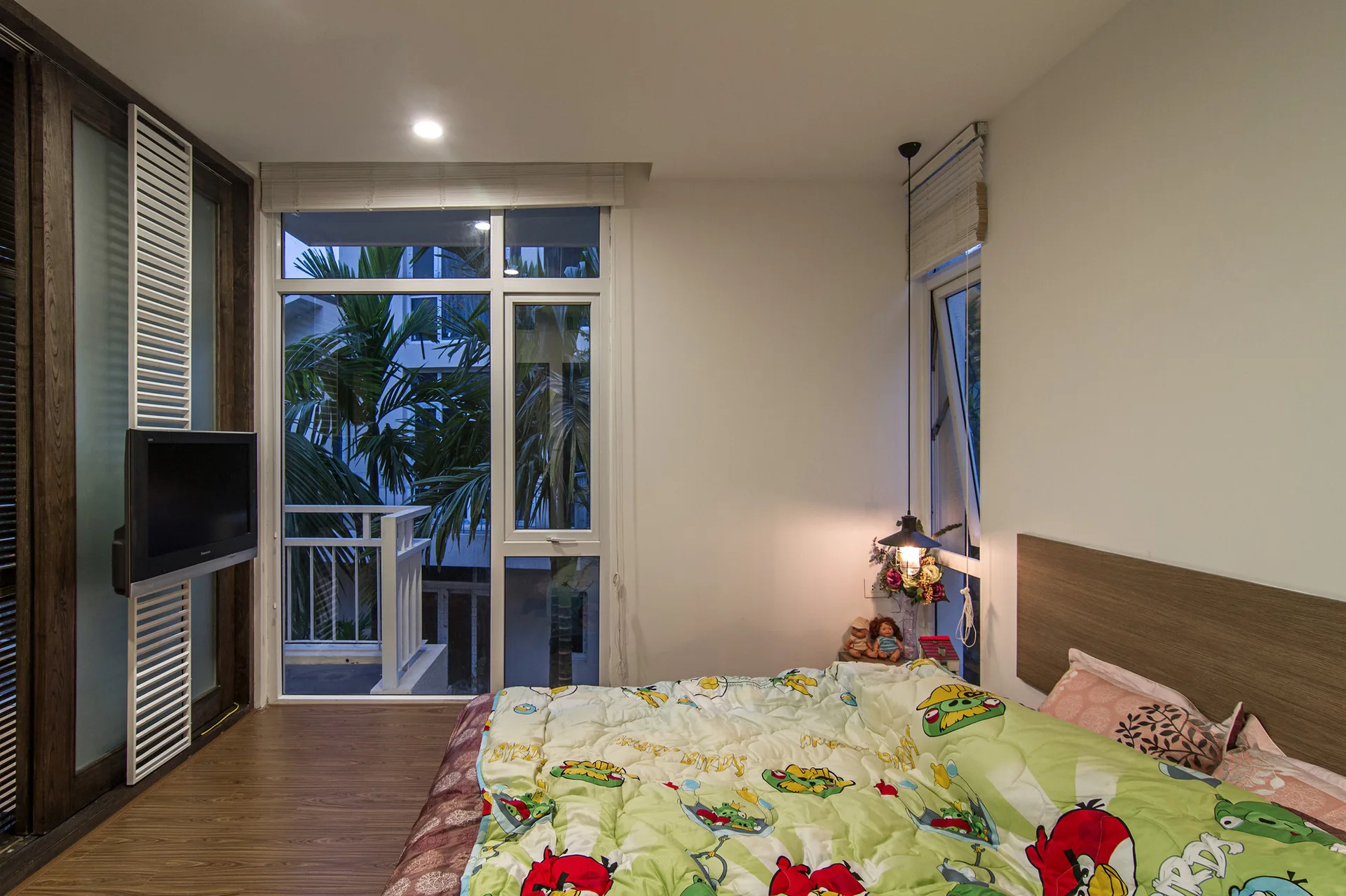 A modern bedroom features a bed with cartoon-print bedding, a wood headboard, a corner pendant light, and a glass door opening to a balcony.