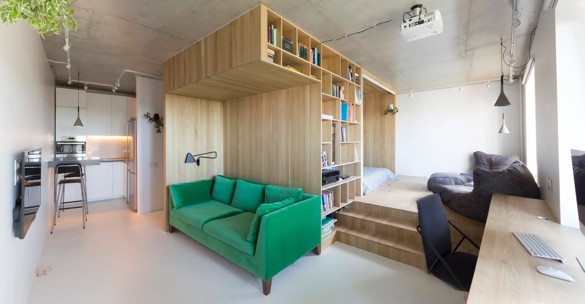A modern studio apartment features an integrated wooden partition unit next to a green velvet sofa and a raised sleeping platform.