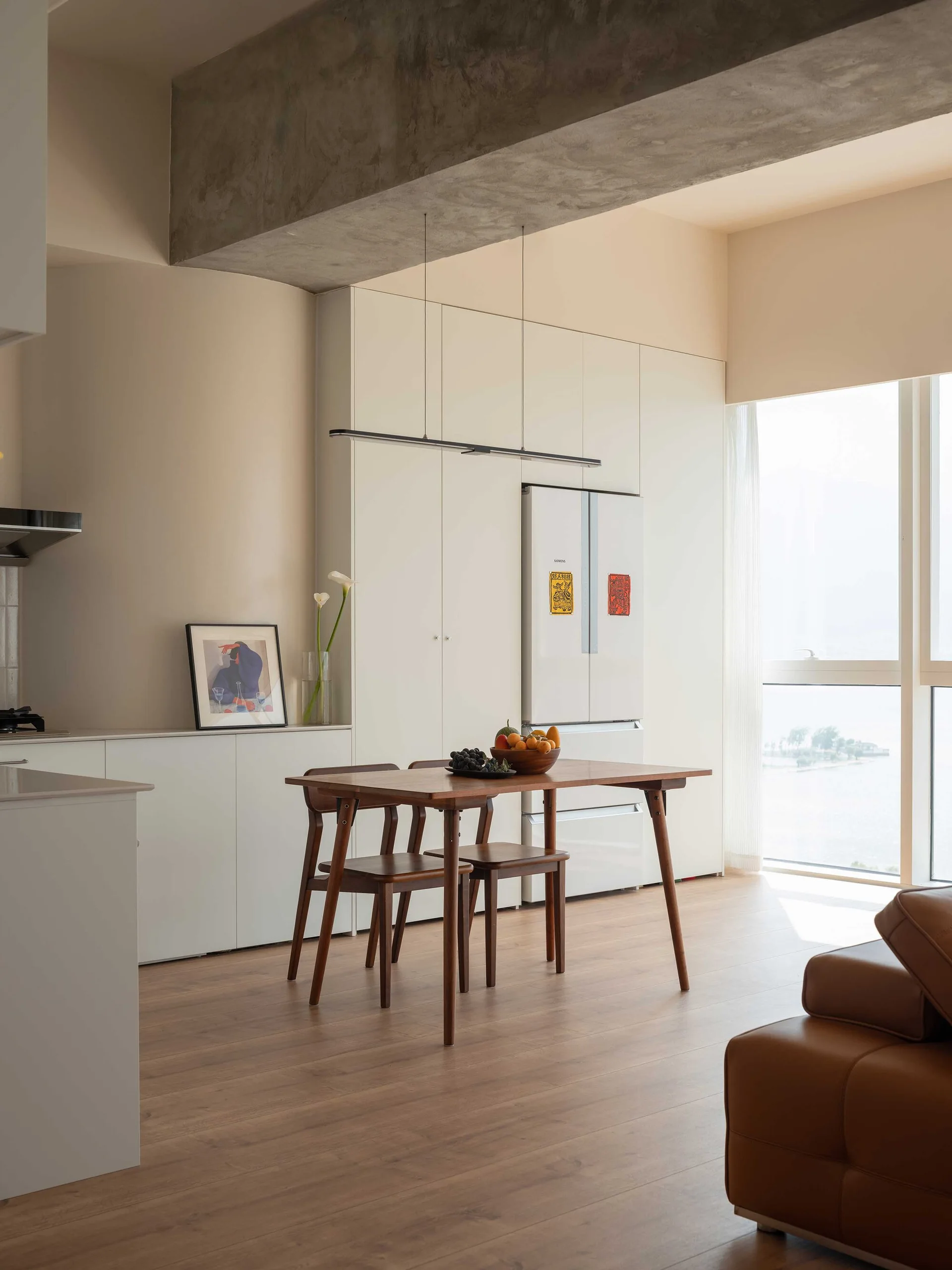 A minimalist dining area featuring a wooden table, mid-century chairs, seamless white cabinetry, and an exposed concrete ceiling beam.