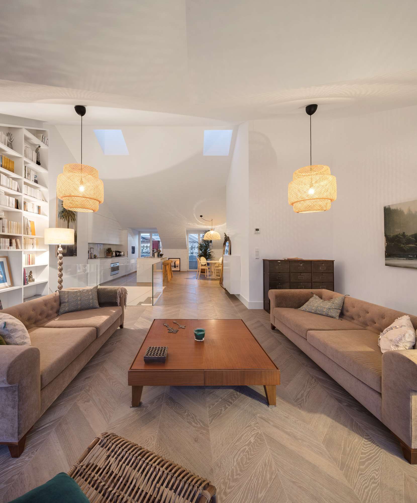A spacious living room features two tufted tan sofas, a central wood coffee table, chevron parquet flooring, and a large built-in bookshelf.