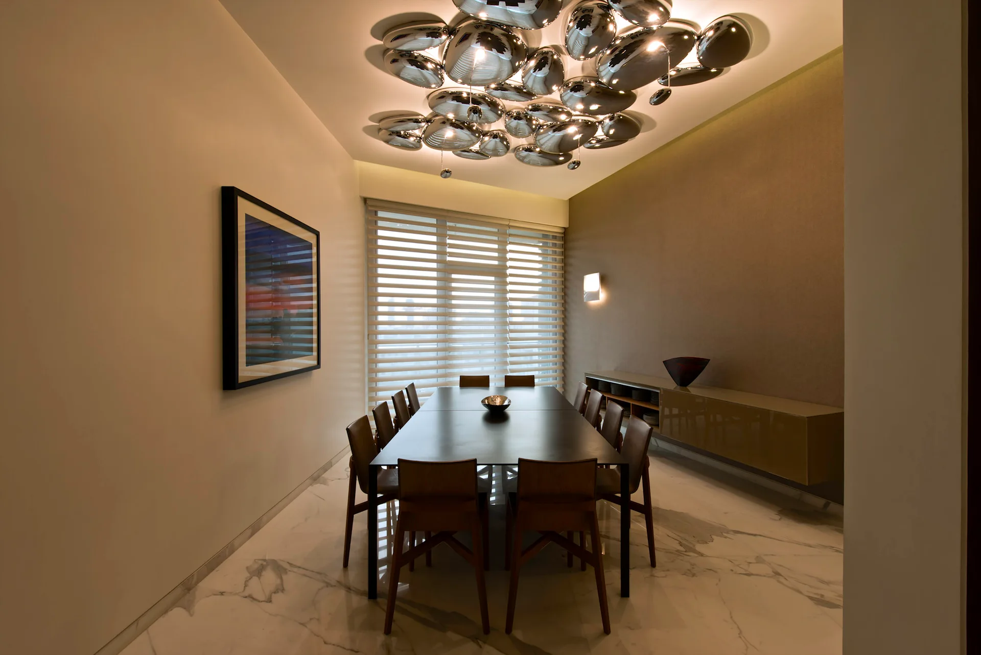 A modern dining room features a dark rectangular table and wooden chairs beneath a large, reflective metallic pebble-style chandelier.
