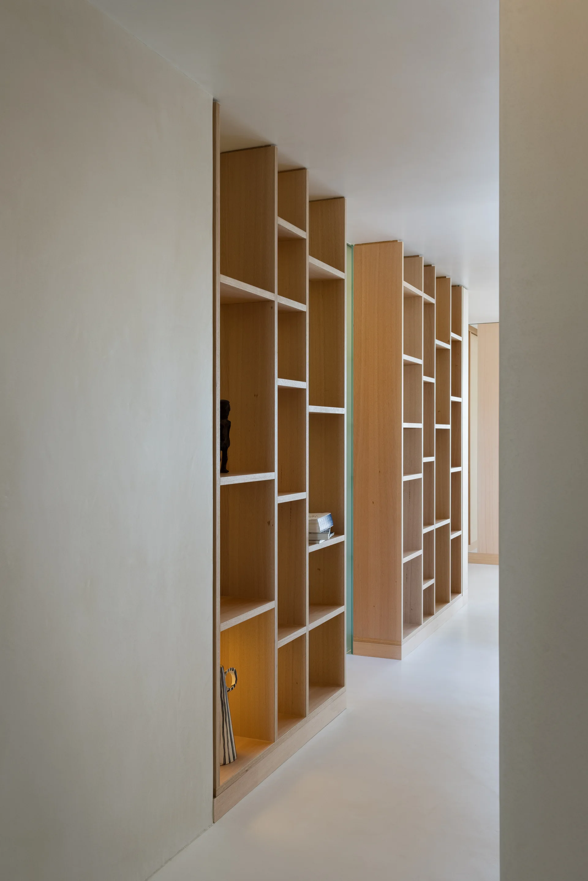 A custom floor-to-ceiling light wood shelving unit with asymmetrical compartments built into a smooth off-white hallway wall.