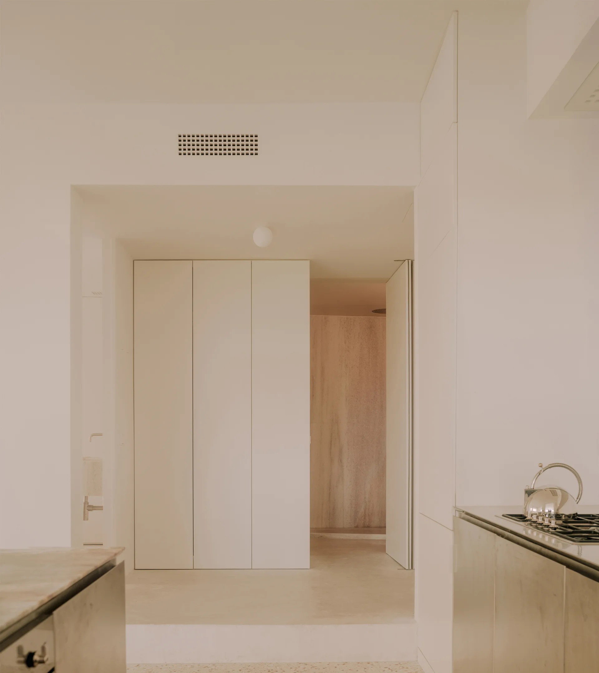 A minimalist kitchen with seamless off-white cabinets, a stainless steel cooktop with a metal kettle, and an open doorway leading to a stone wall.