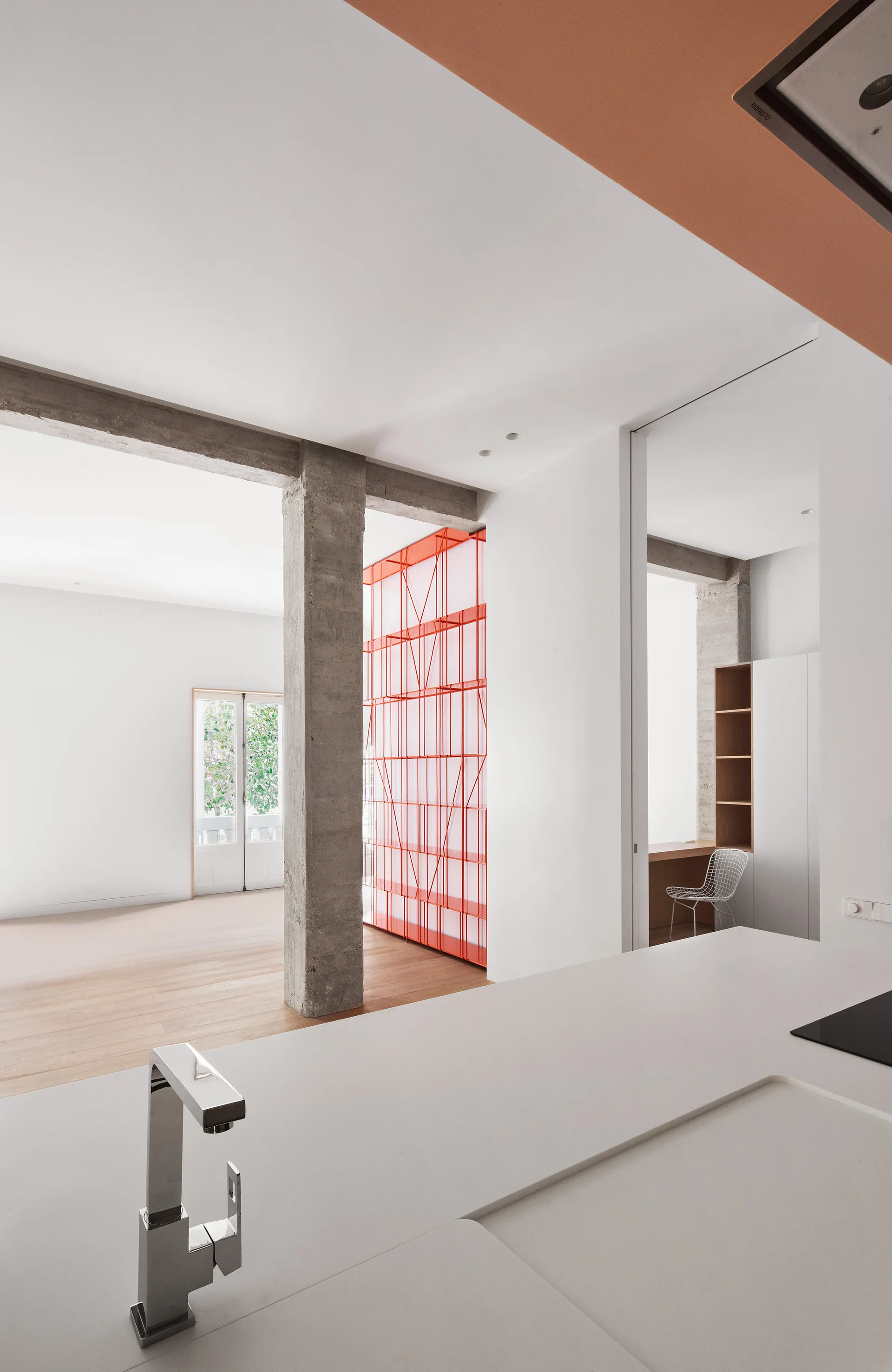 An open-plan kitchen with a white countertop and faucet features an exposed concrete column and a bright red grid shelving partition in the background.