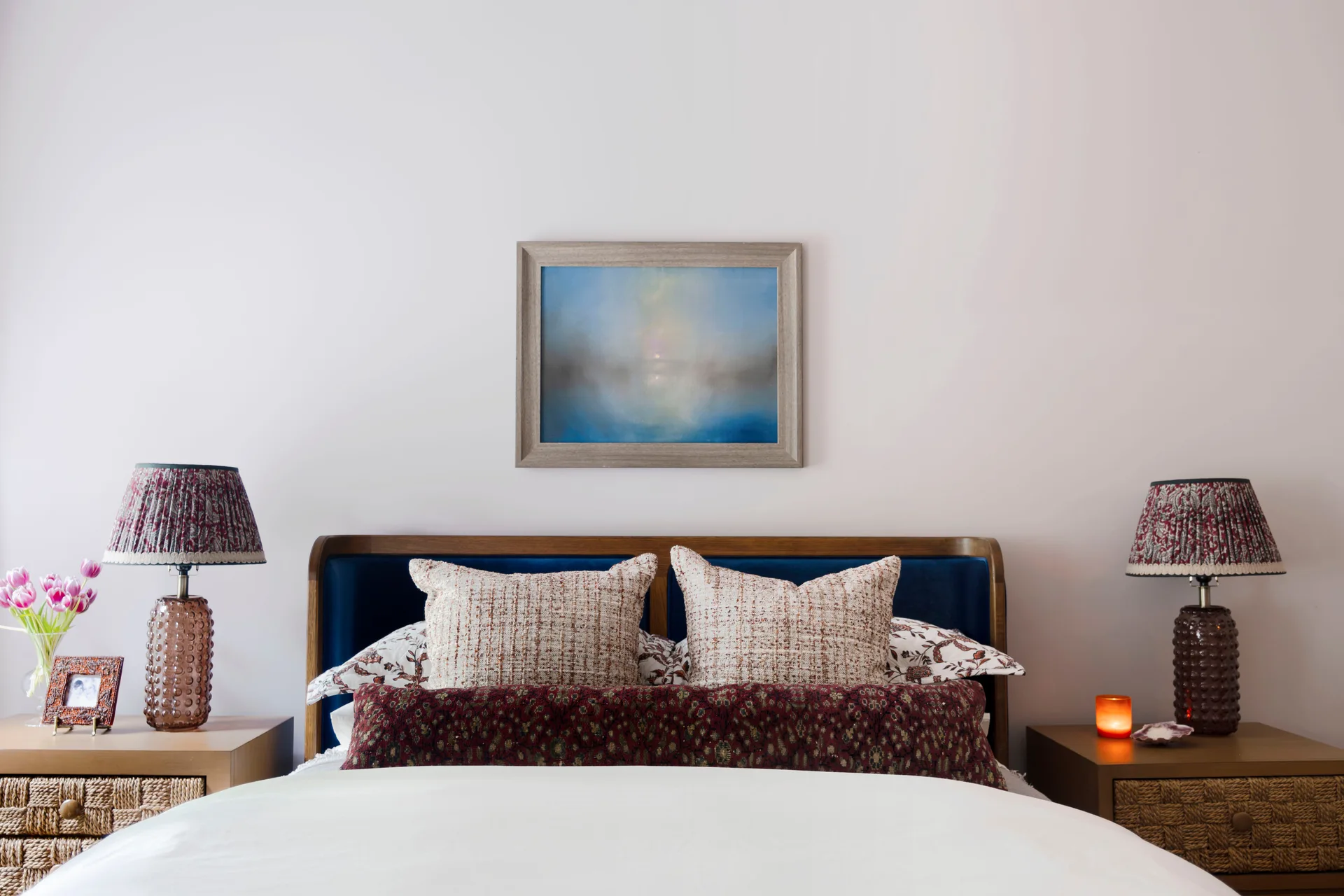 A symmetrical bedroom features a blue velvet headboard, wood nightstands with woven drawers, and patterned lamps flanking the bed.