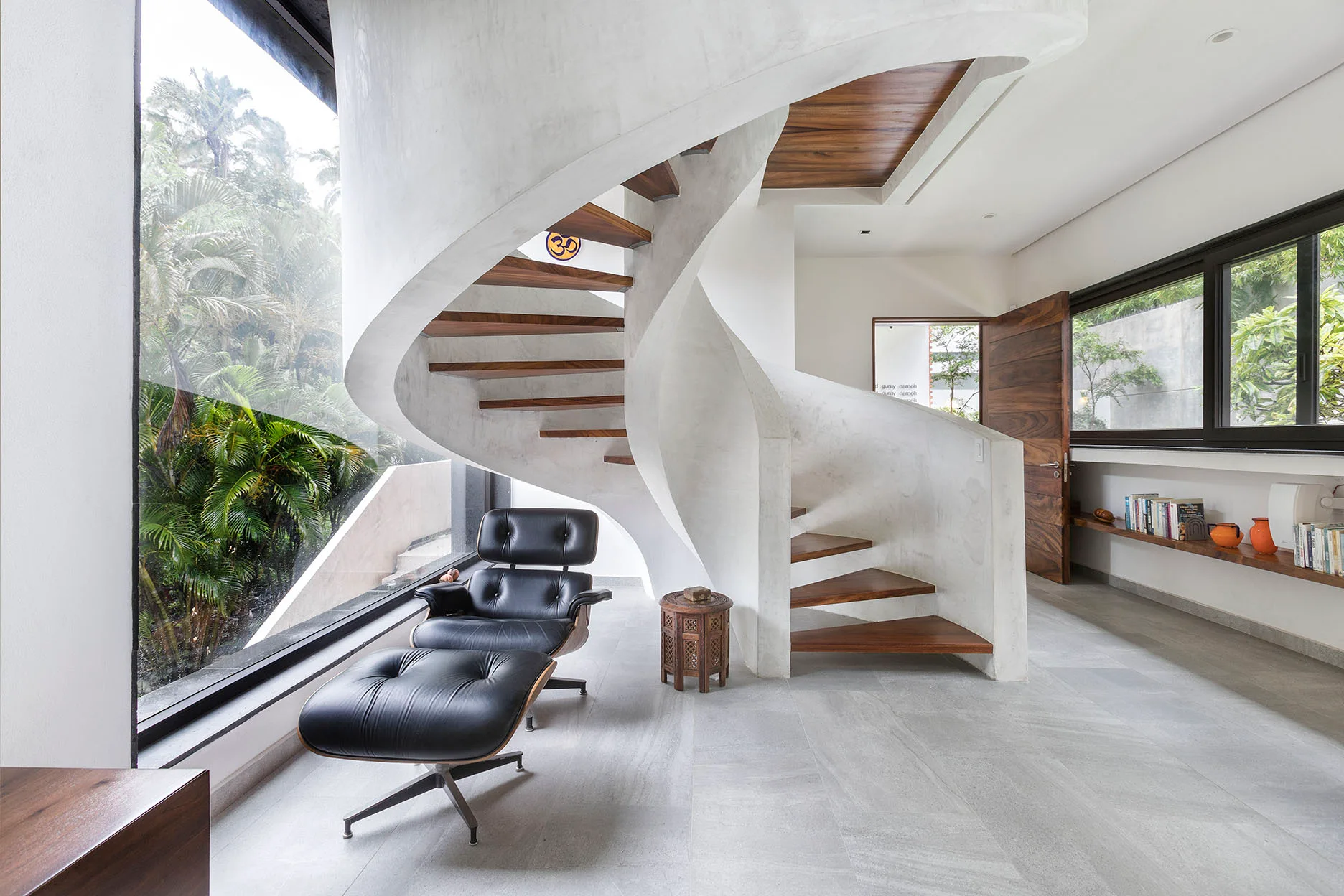 A modern room features a concrete spiral staircase with wood steps next to a black lounge chair and a large window looking out at green trees.