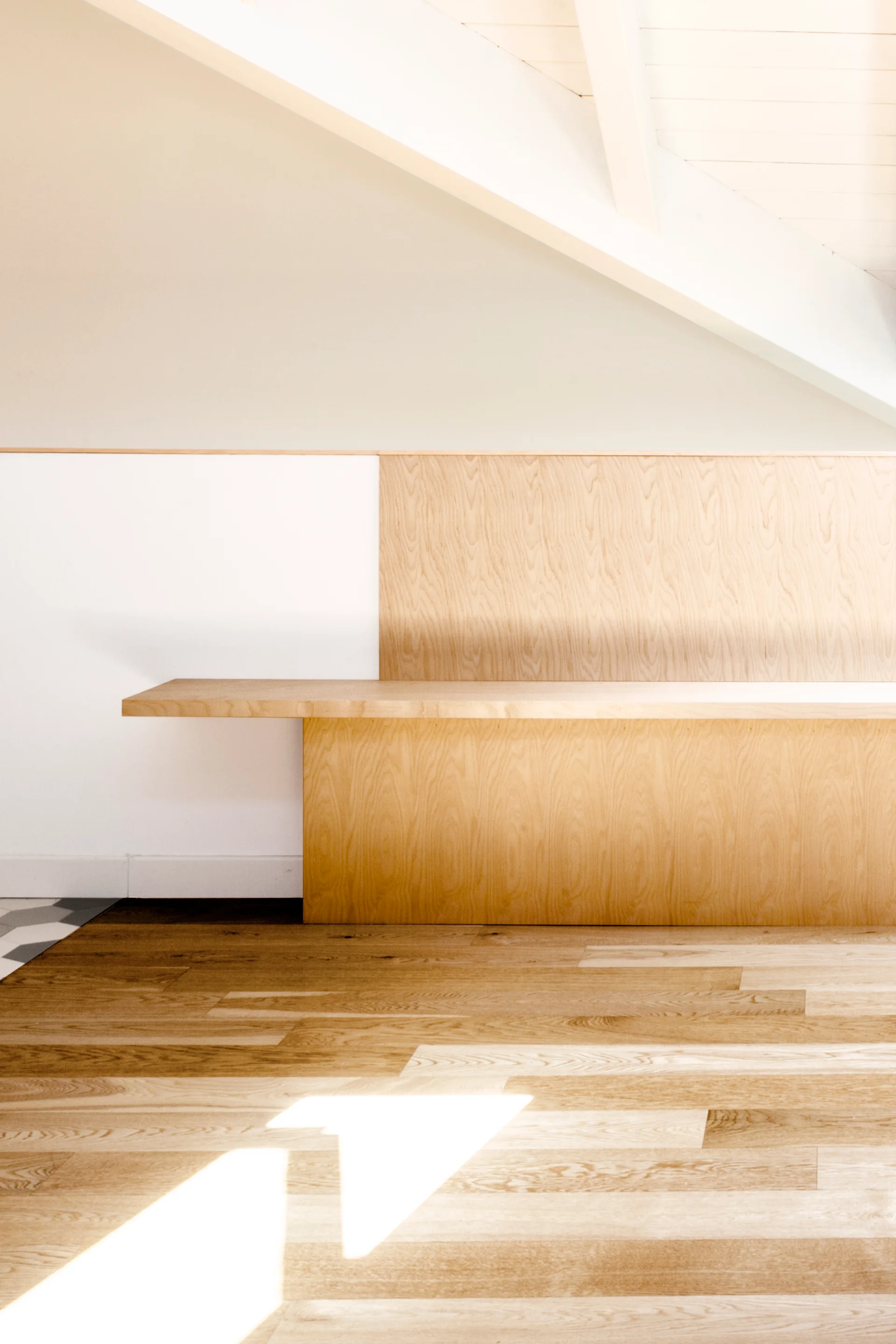 A built-in light wood bench is integrated into a paneled wall beneath a slanted white ceiling with a structural beam.