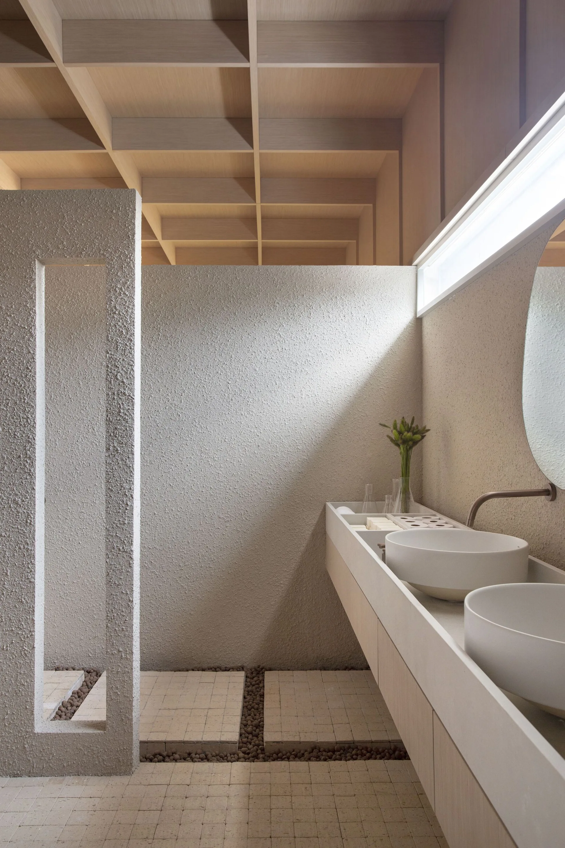 A minimalist bathroom featuring a light wood grid ceiling, dual white vessel sinks on a beige vanity, and heavily textured sand-colored plaster walls.