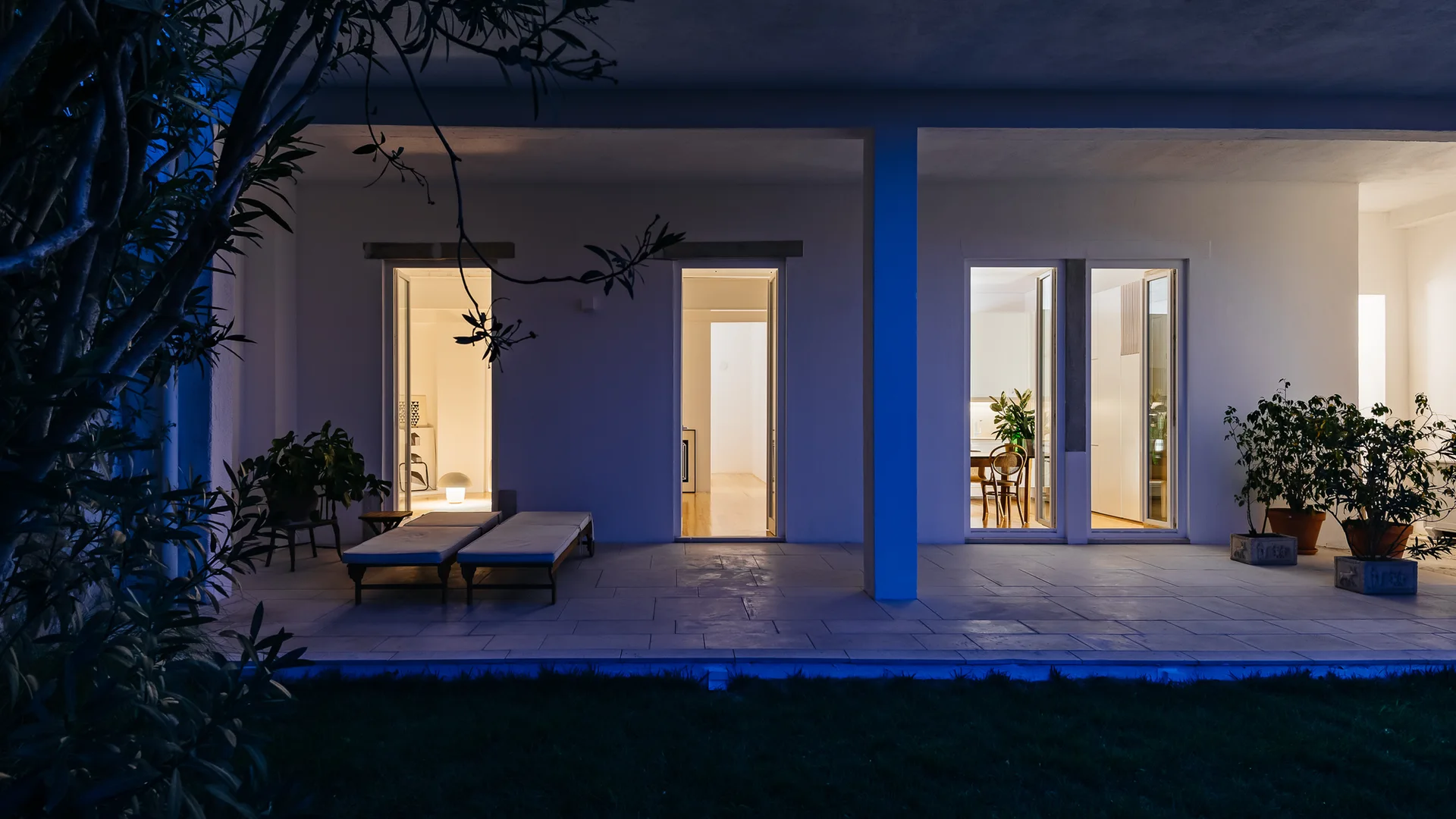 A covered stone patio at night features two lounge chairs beside tall glass doors revealing warm lit interior rooms.