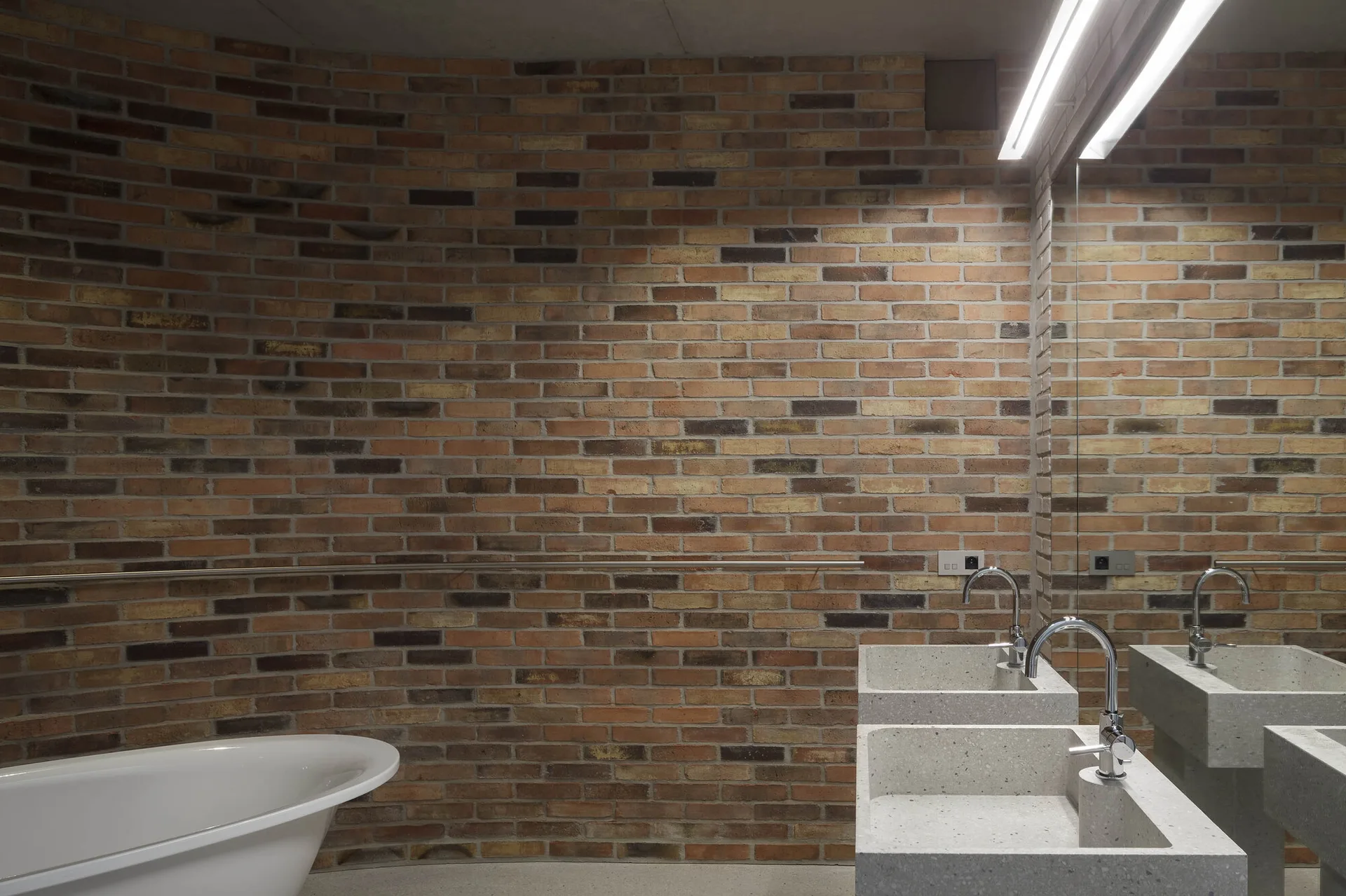 A modern bathroom featuring a curved exposed brick wall, a freestanding white bathtub, and a double concrete-style vanity with chrome faucets.