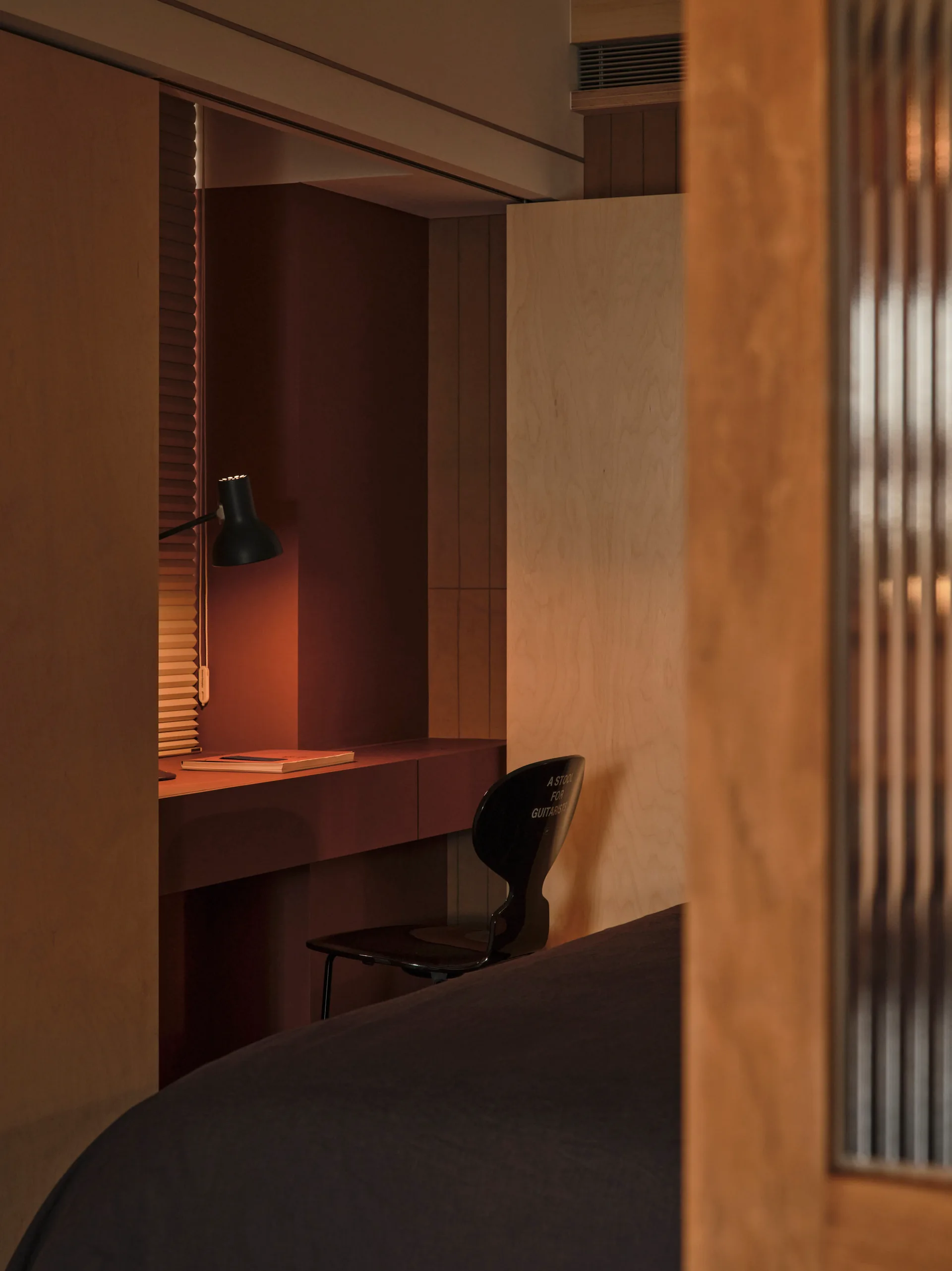 A dimly lit bedroom alcove features a built-in deep red desk with a black task lamp and a black chair against light wood paneled walls.