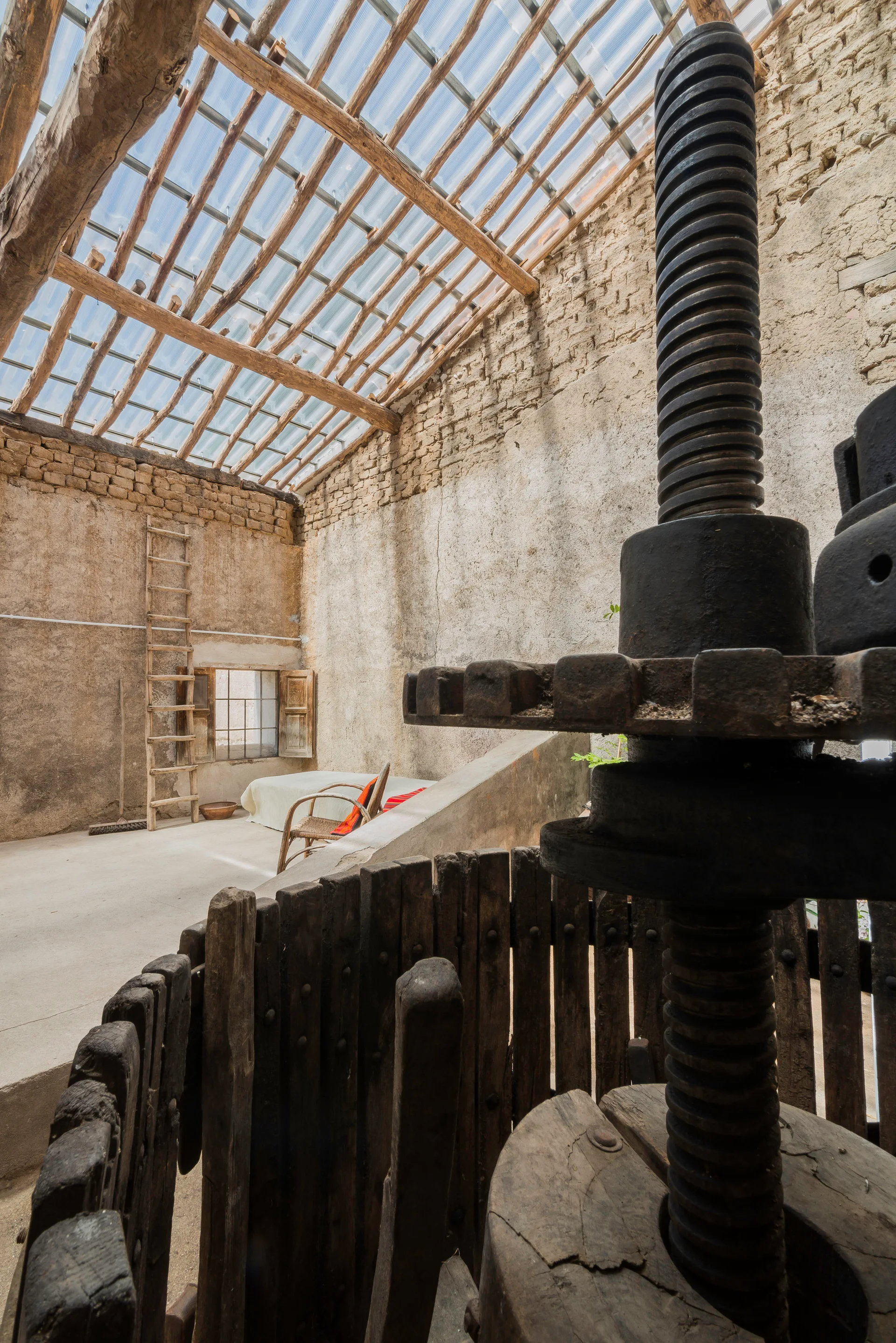 A rustic barn interior featuring a large vintage wine press, coarse plaster walls, a wooden ladder, and a translucent corrugated roof.