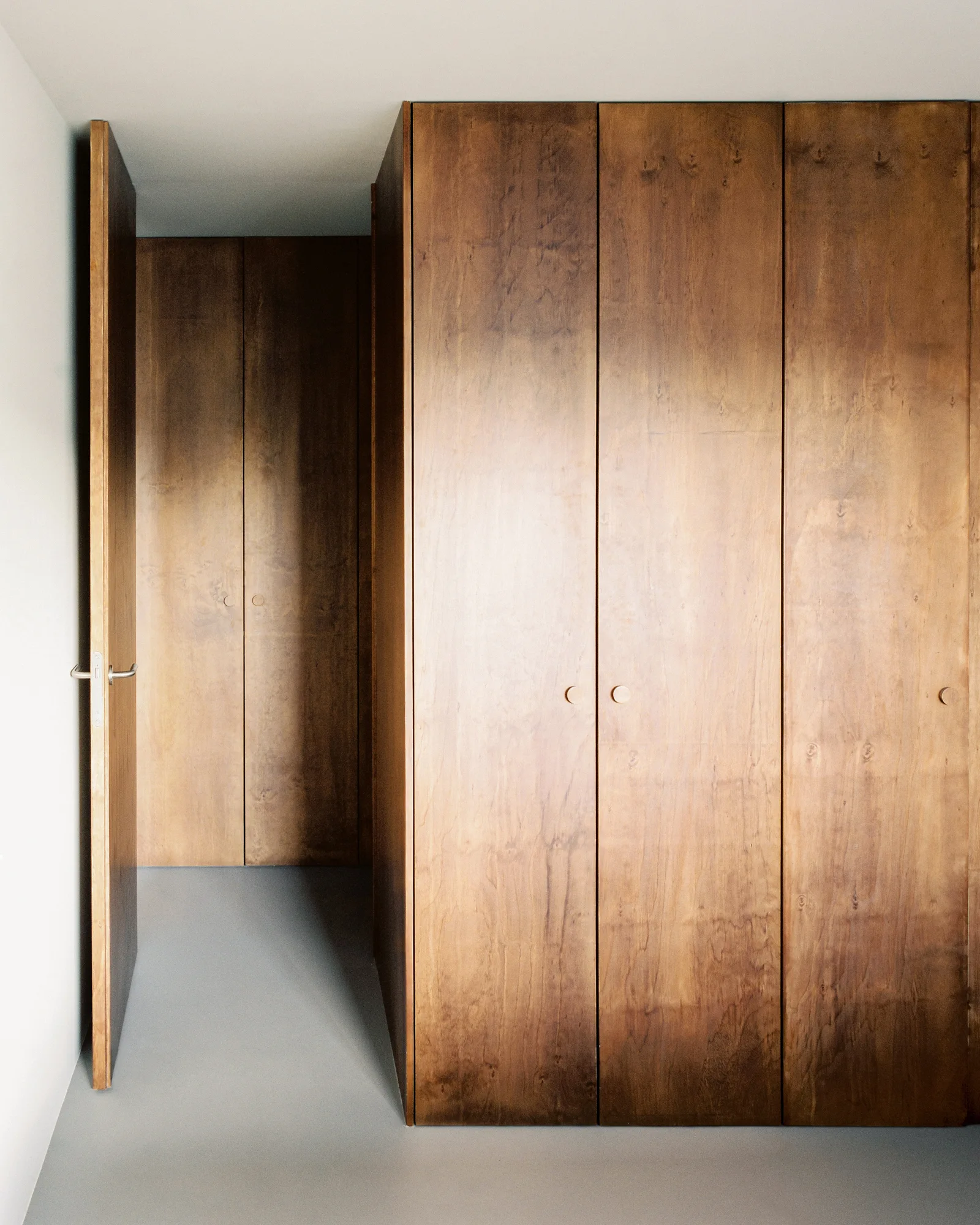 A minimalist hallway featuring floor to ceiling wood wardrobe cabinets, a matching open door, and a smooth grey floor.