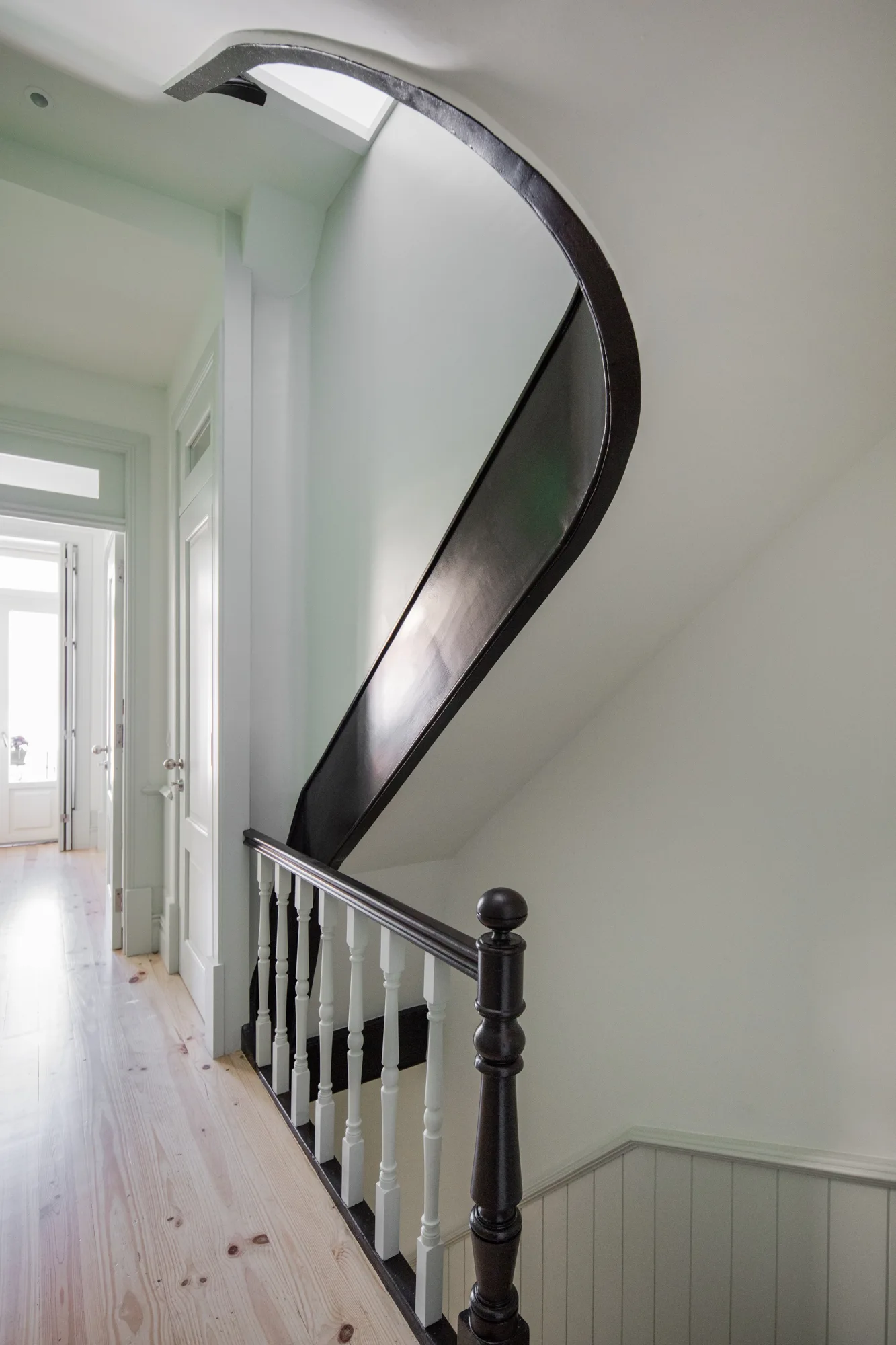 A staircase with a dramatic curved black handrail stands next to white balusters, pale green walls, and light wood flooring in a bright hallway.