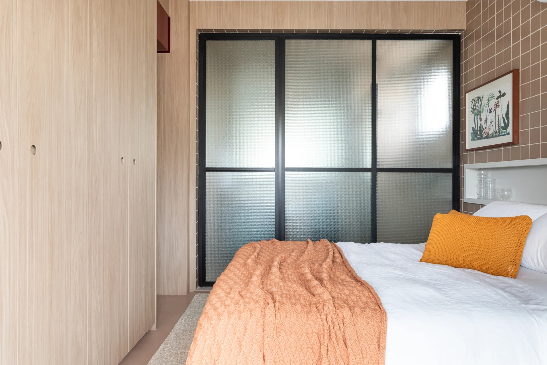 A modern bedroom features a light wood wardrobe, a bed with an orange throw, and a black-framed frosted glass sliding partition wall.