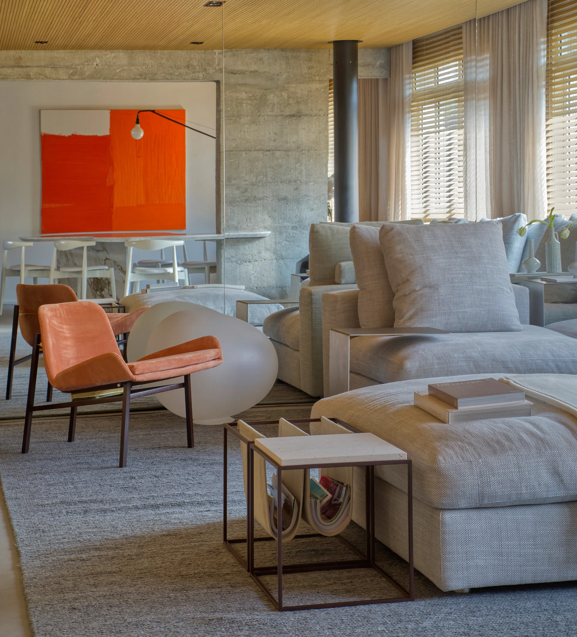 A modern living room featuring a grey sectional, orange accent chairs, exposed concrete walls, and a warm slatted wood ceiling.