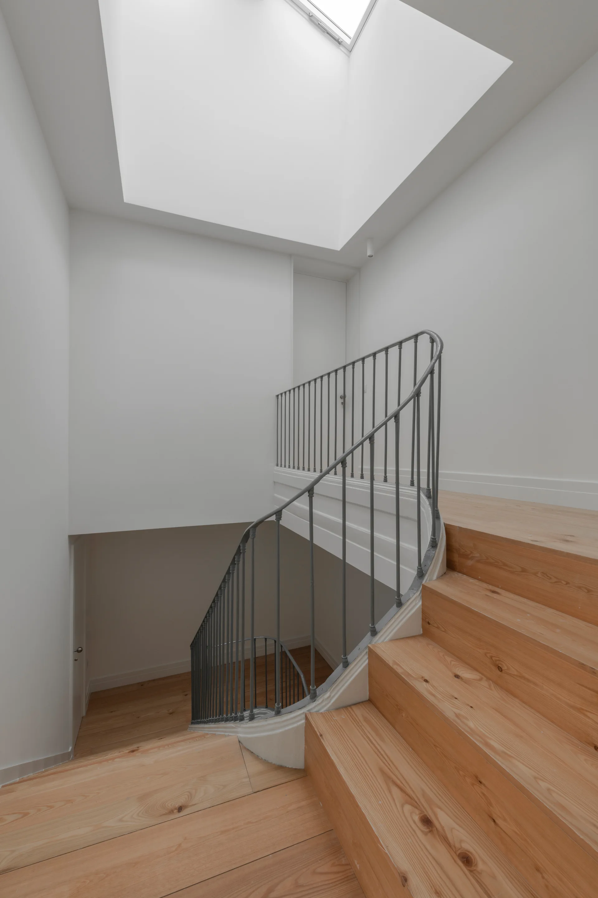 A minimalist wooden staircase winding down with a slender grey iron railing under a large square ceiling skylight.