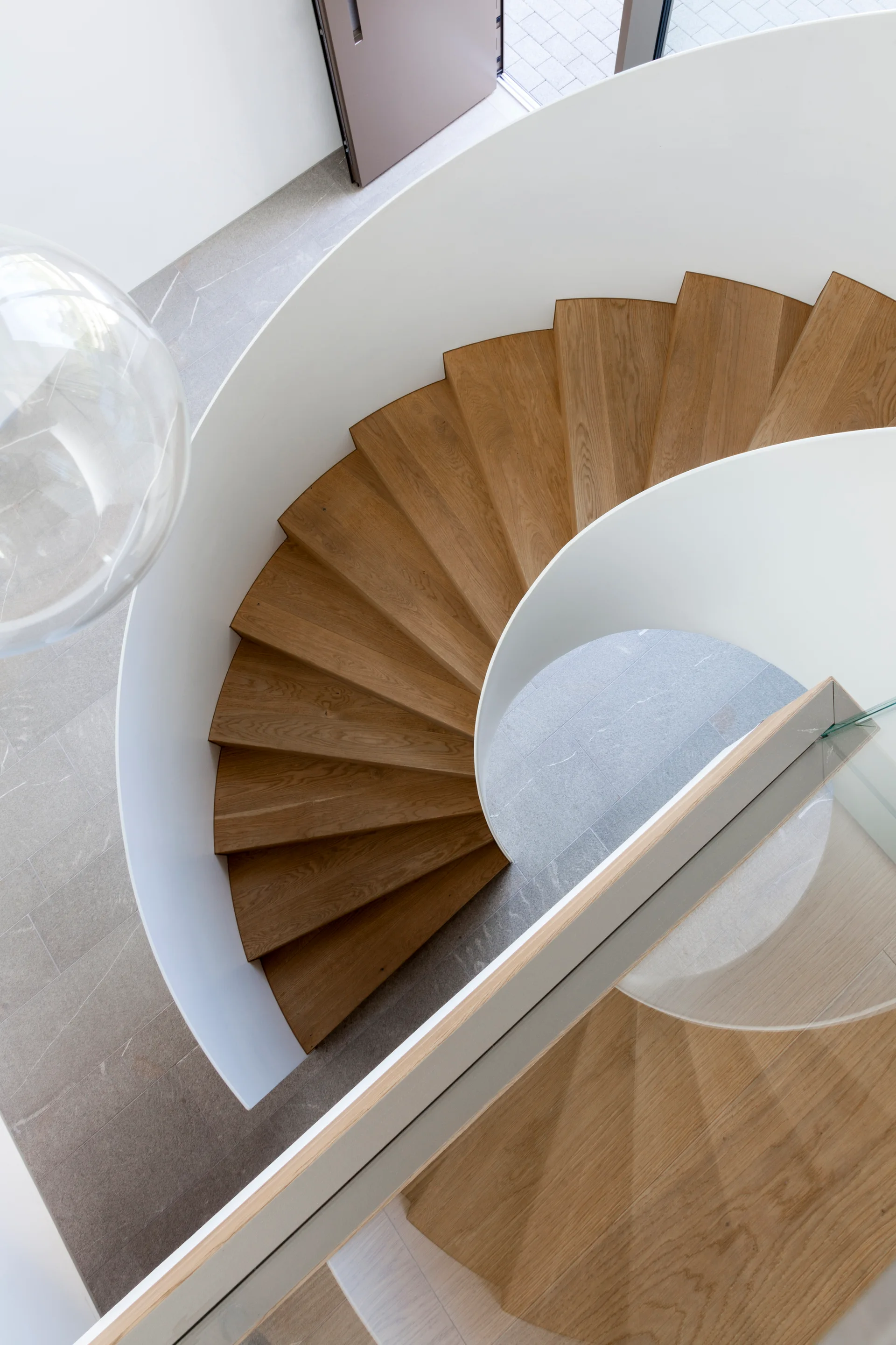 Overhead view of a spiral staircase featuring medium-tone wood treads, a curved white outer structure, grey stone tile flooring, and a glass railing.