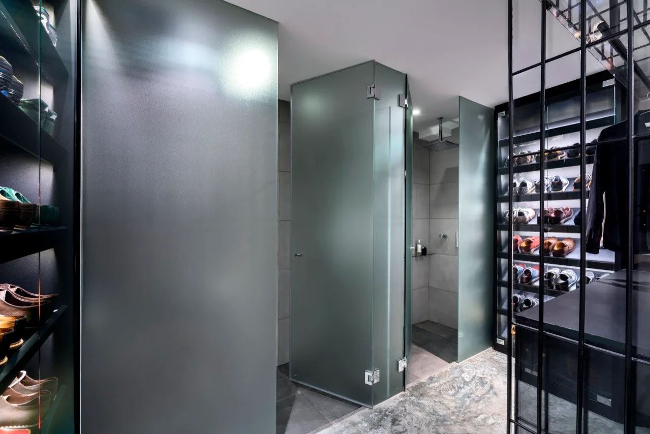 A modern dressing room features integrated black metal shoe shelving with built-in backlighting next to a frosted glass shower enclosure.