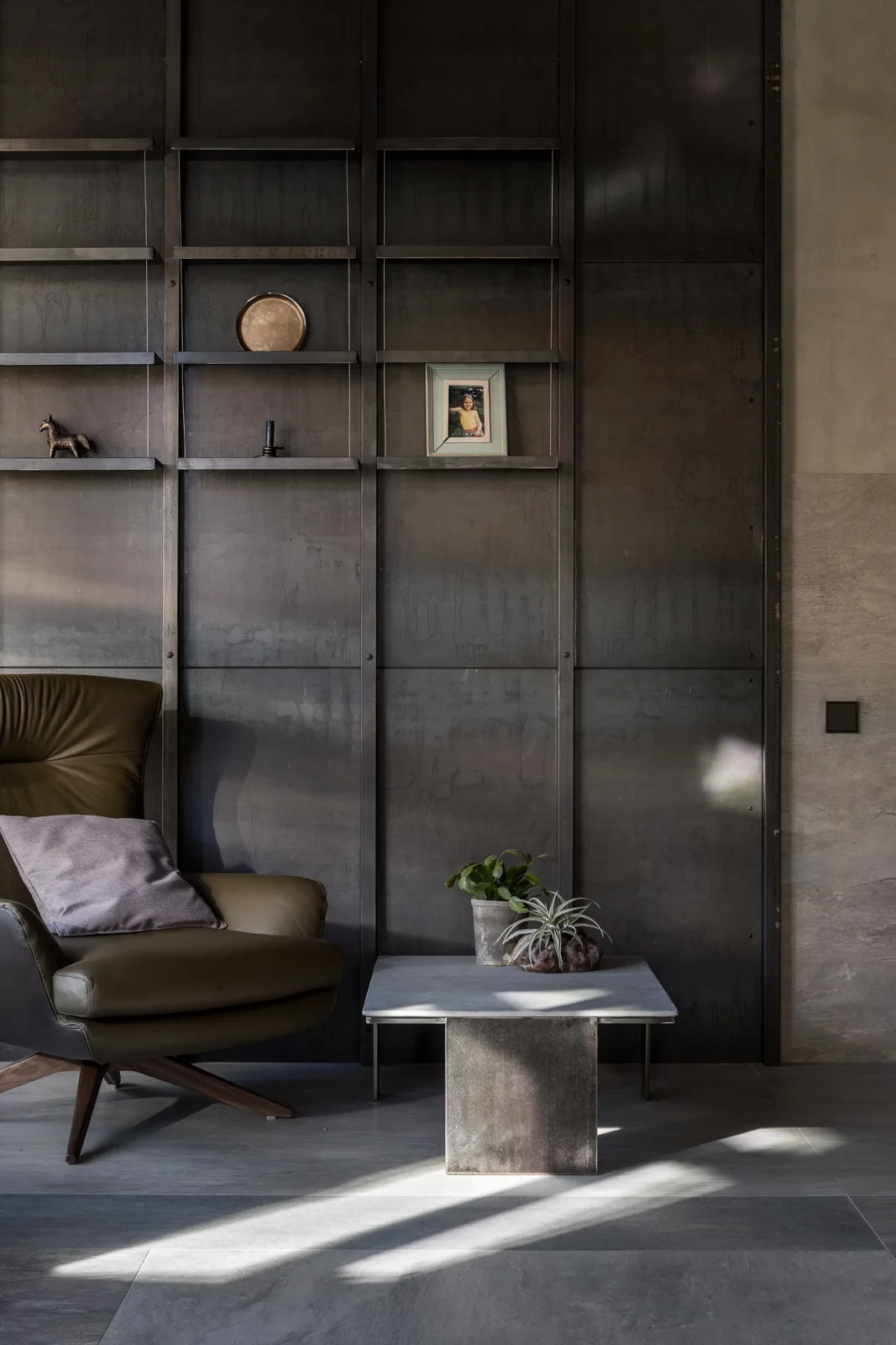 An olive green leather armchair stands next to a low stone-topped table against a dark grey metal paneled wall with integrated shelves.