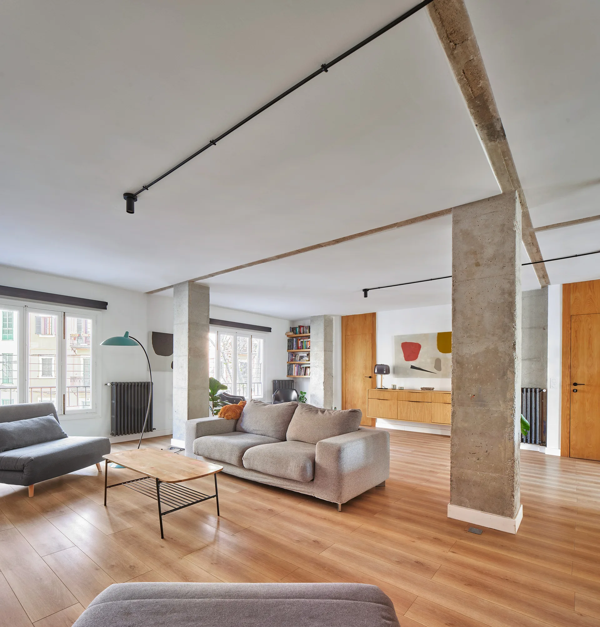 An open plan living room features a grey sofa, a wood coffee table, two exposed concrete columns, and natural wood flooring.