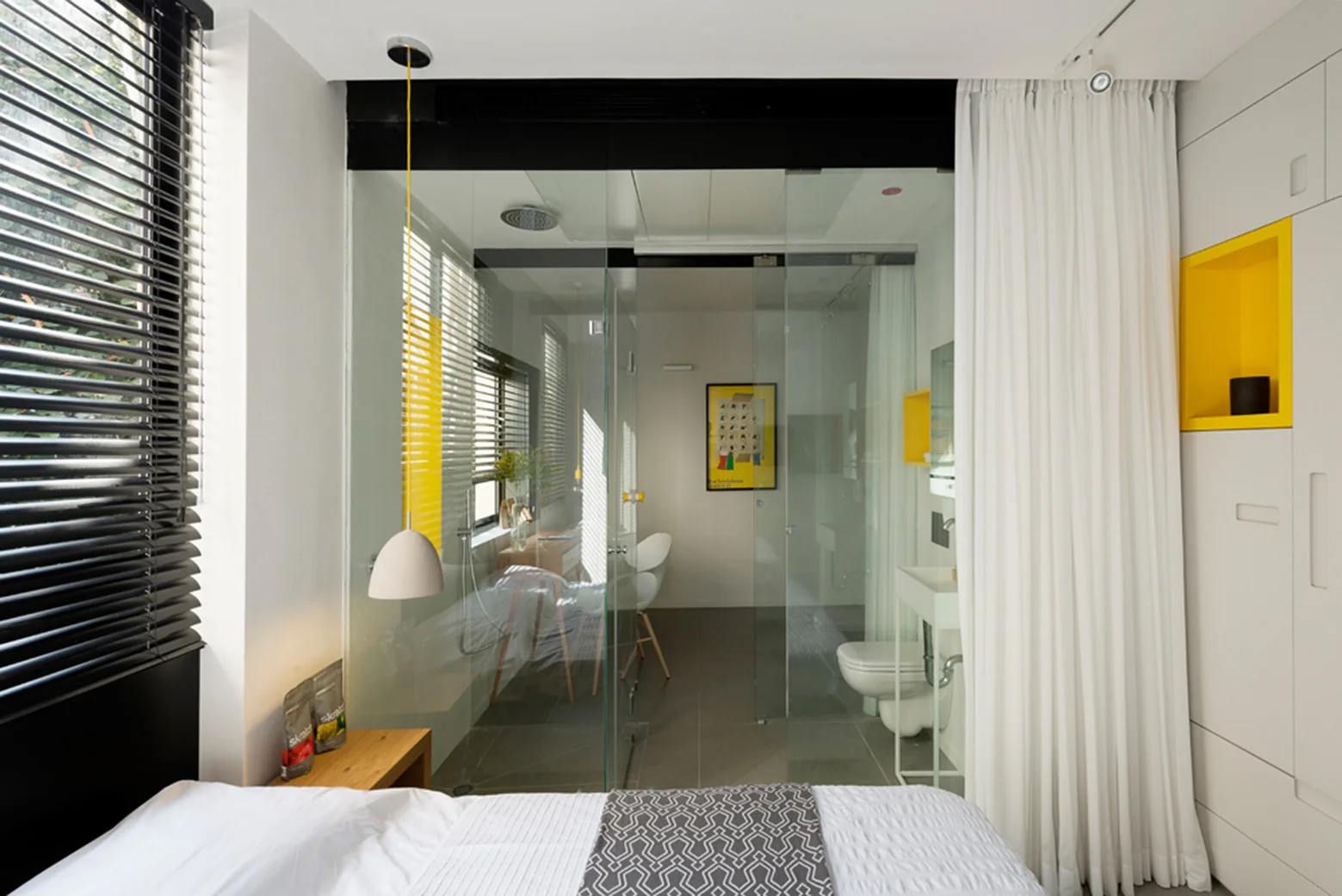 A modern bedroom features a glass-enclosed bathroom, a built-in wardrobe with a yellow alcove, white curtains, and a bed in the foreground.