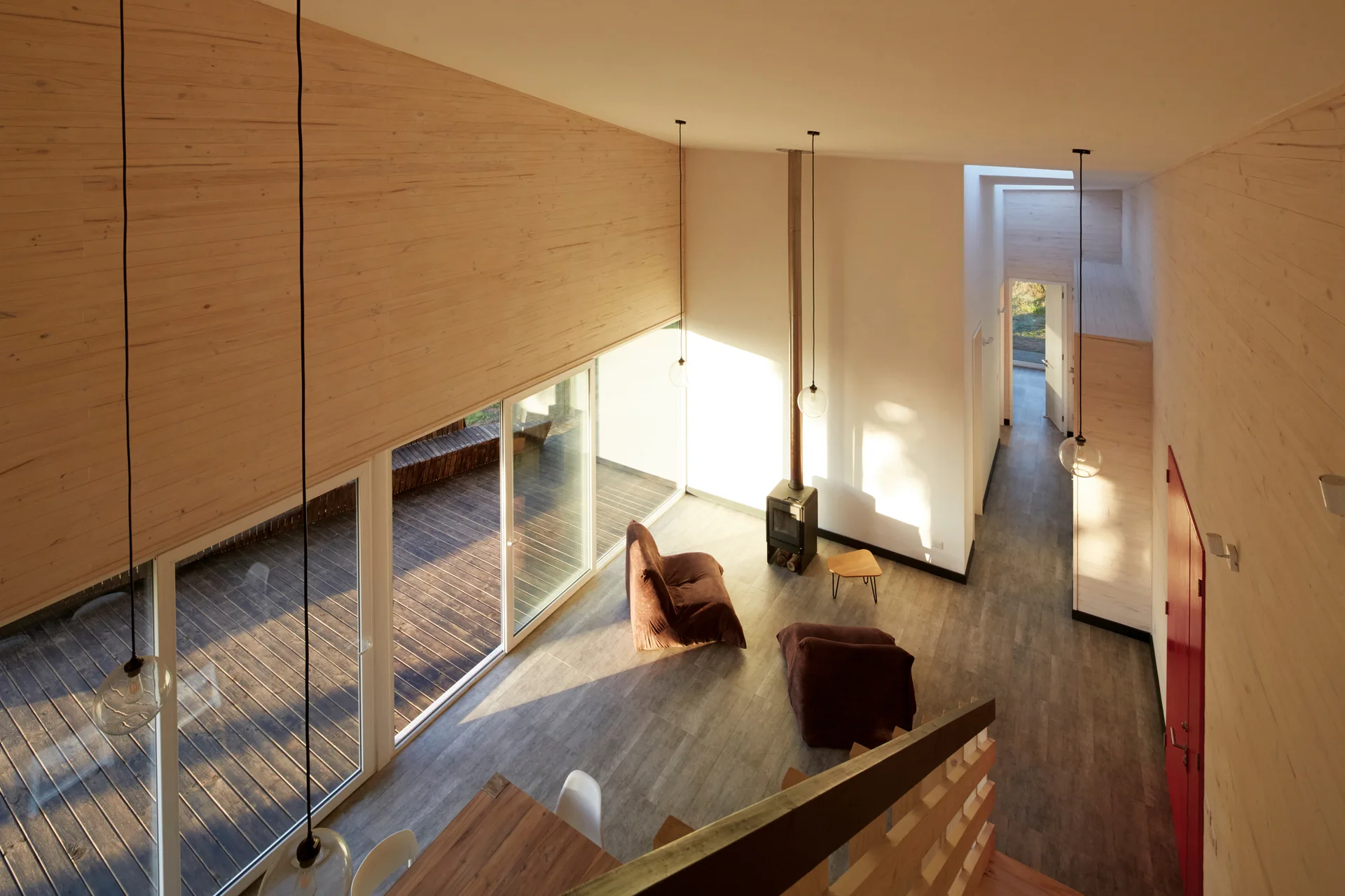 A high-angle view of a minimalist living room featuring light wood paneled walls, sliding glass doors to a deck, and low-profile brown floor chairs.