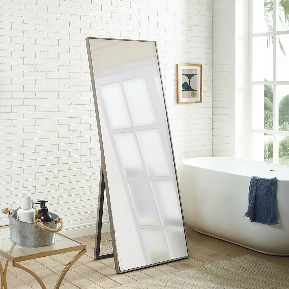 A tall rectangular floor mirror with a thin dark metal frame stands on a wooden floor in a bathroom with a white tub and brick wall.